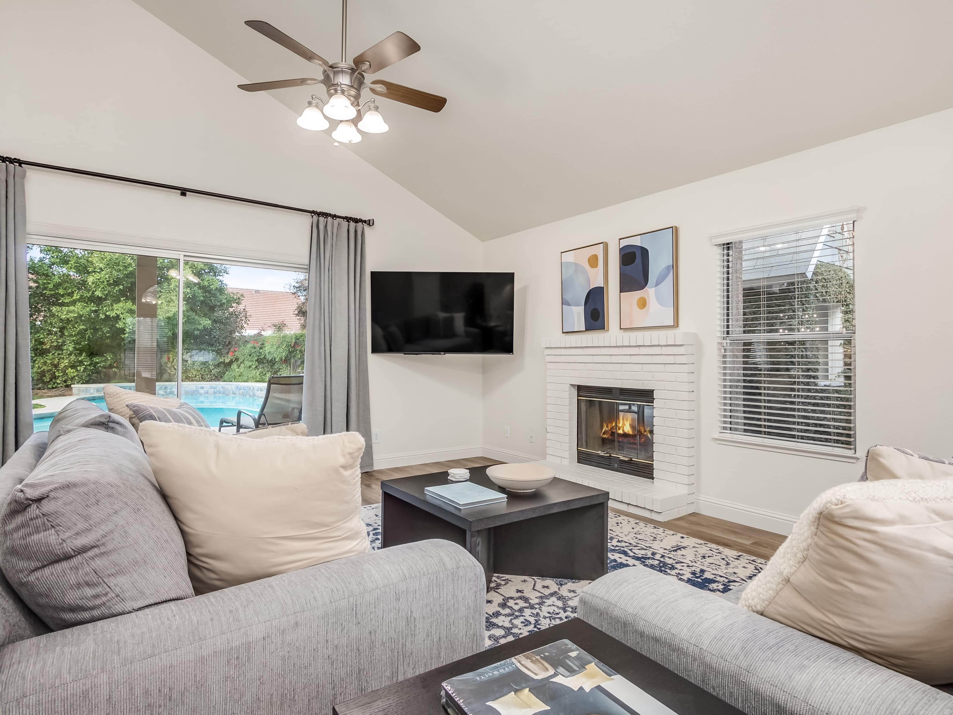 A living room features two gray sofas facing a coffee table, a fireplace, a large TV mounted on the wall, and a pool visible through large windows.