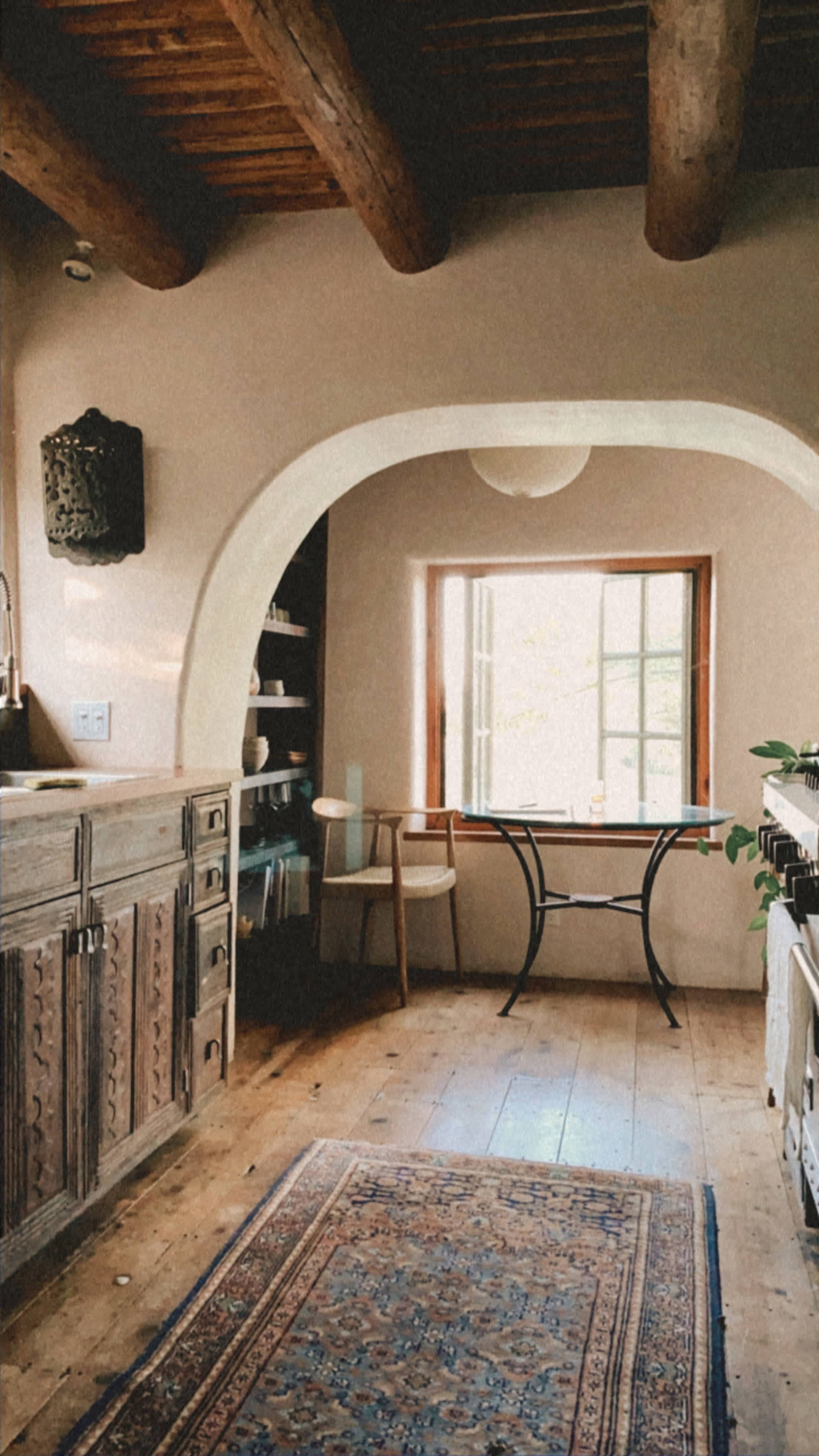 A rustic kitchen features wooden beams, a curved archway, a central table, and a window allowing natural light to enter.