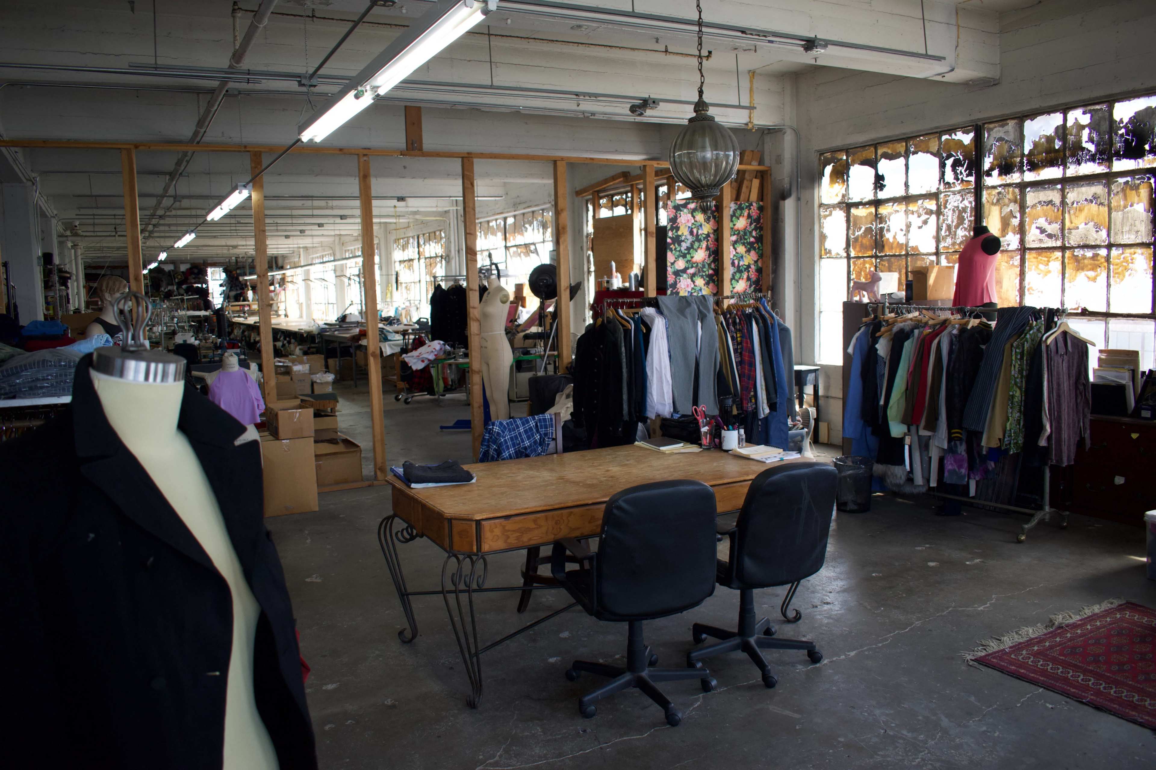 The image shows a spacious garment workshop with clothing racks, a wooden table, and mannequins, surrounded by large windows.