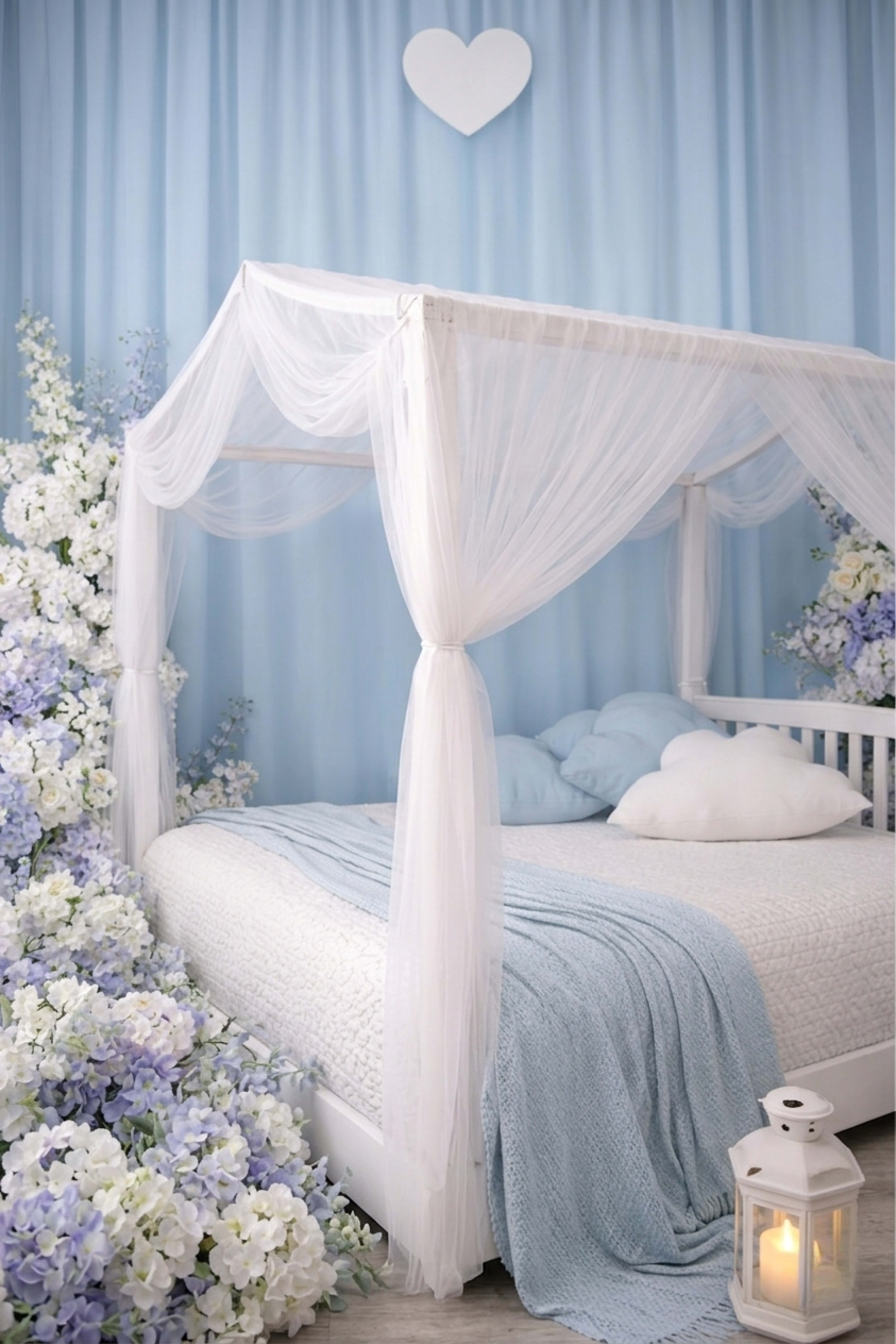A white canopy bed is decorated with light blue bedding, surrounded by artificial flowers and a lantern on the floor.