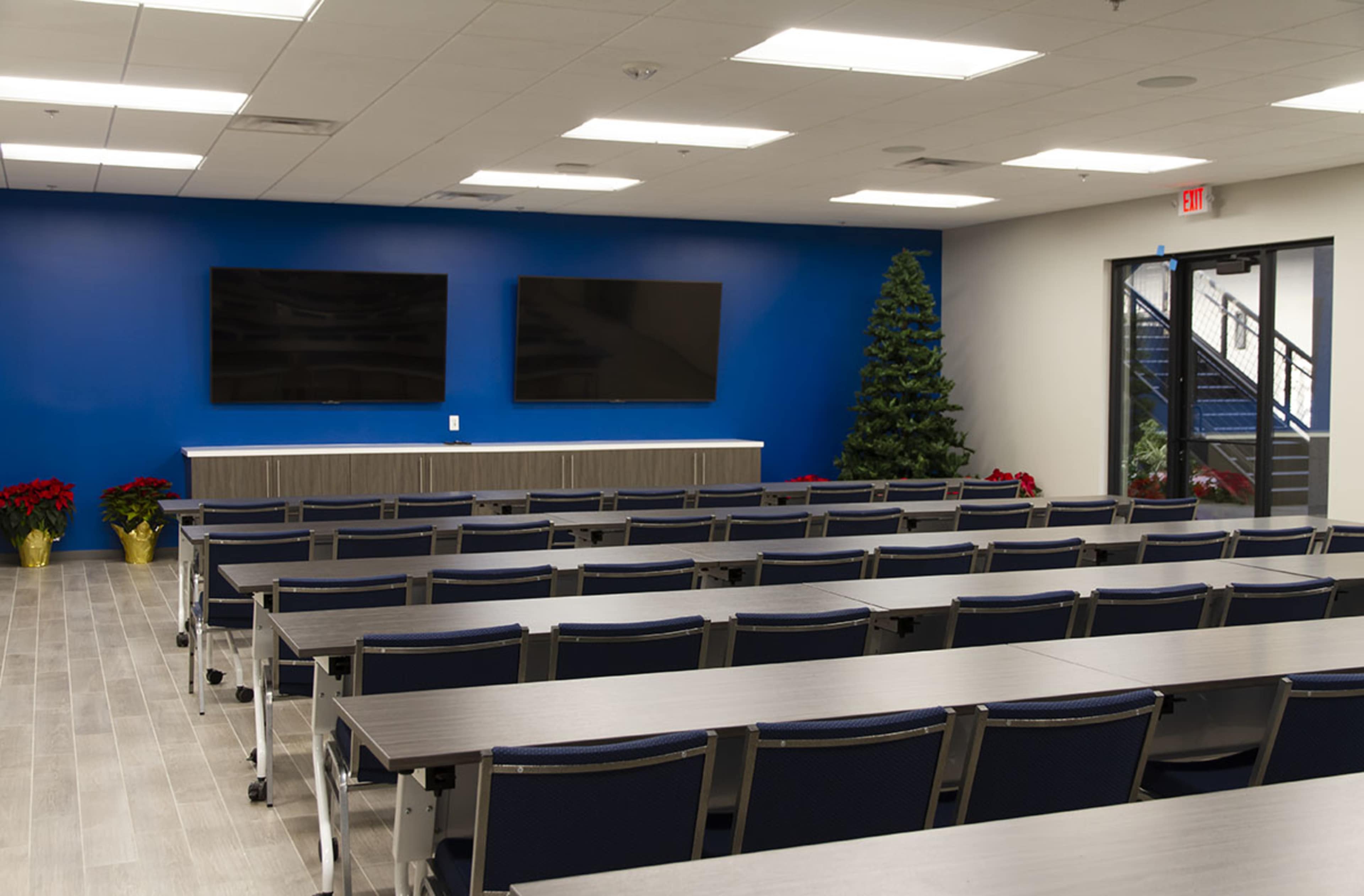 A modern conference room with rows of tables and chairs facing two wall-mounted screens and a decorative tree in the corner.