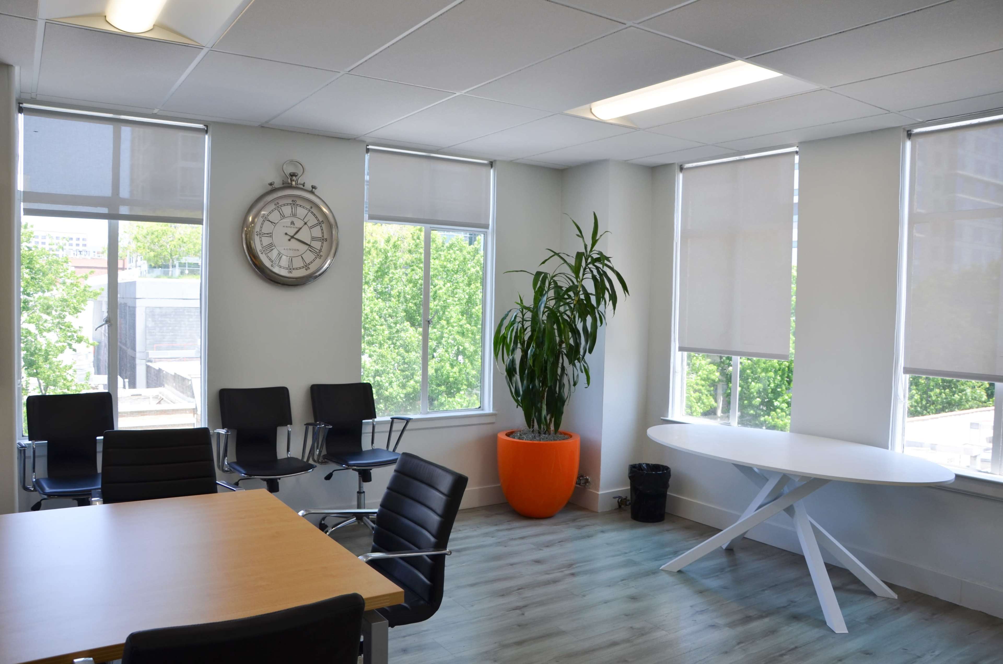 The image shows a modern office space with a round white table, black chairs, a large wall clock, a potted plant, and windows allowing natural light.
