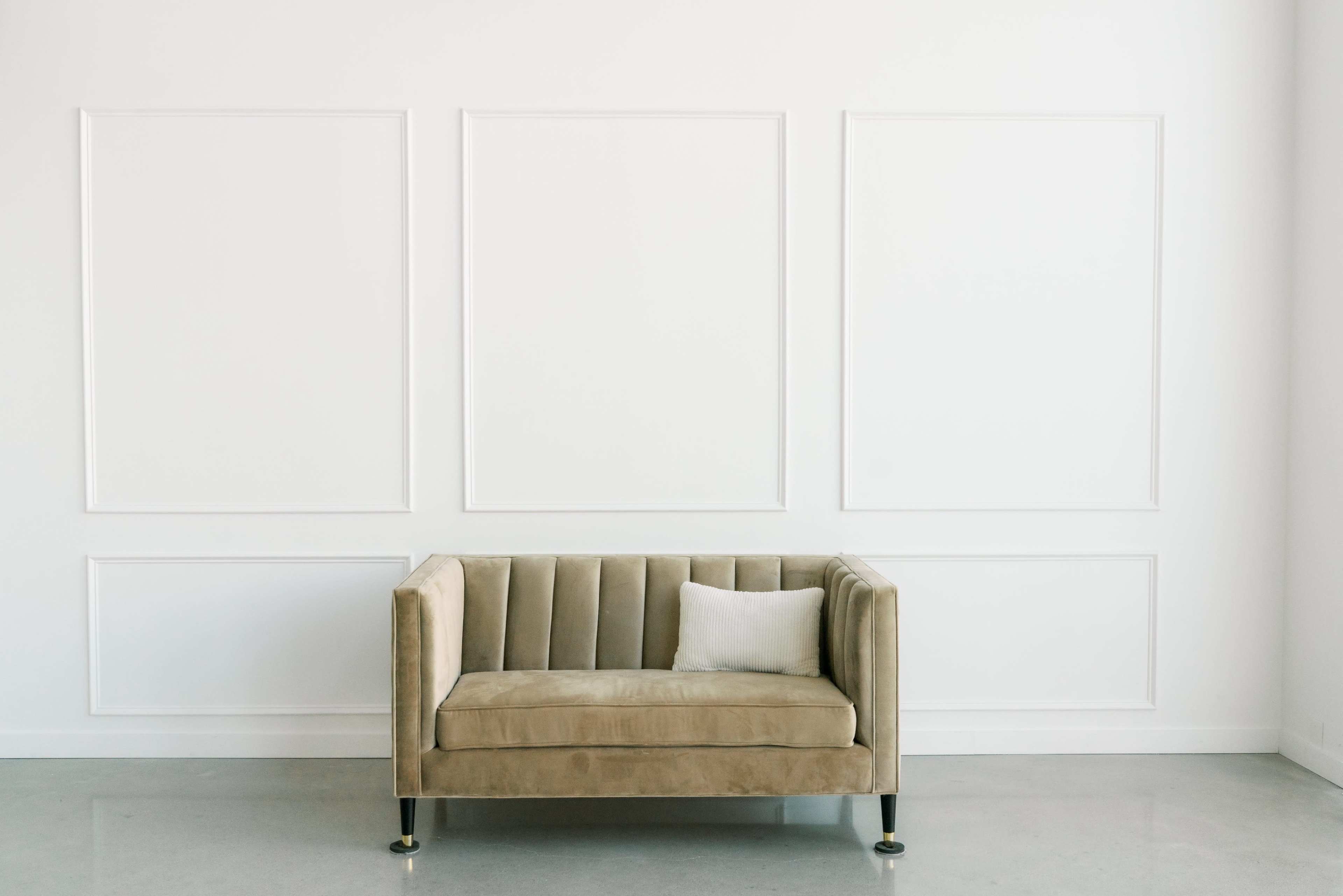 A beige velvet couch sits against a plain white wall with three plain frames outlined in molding.