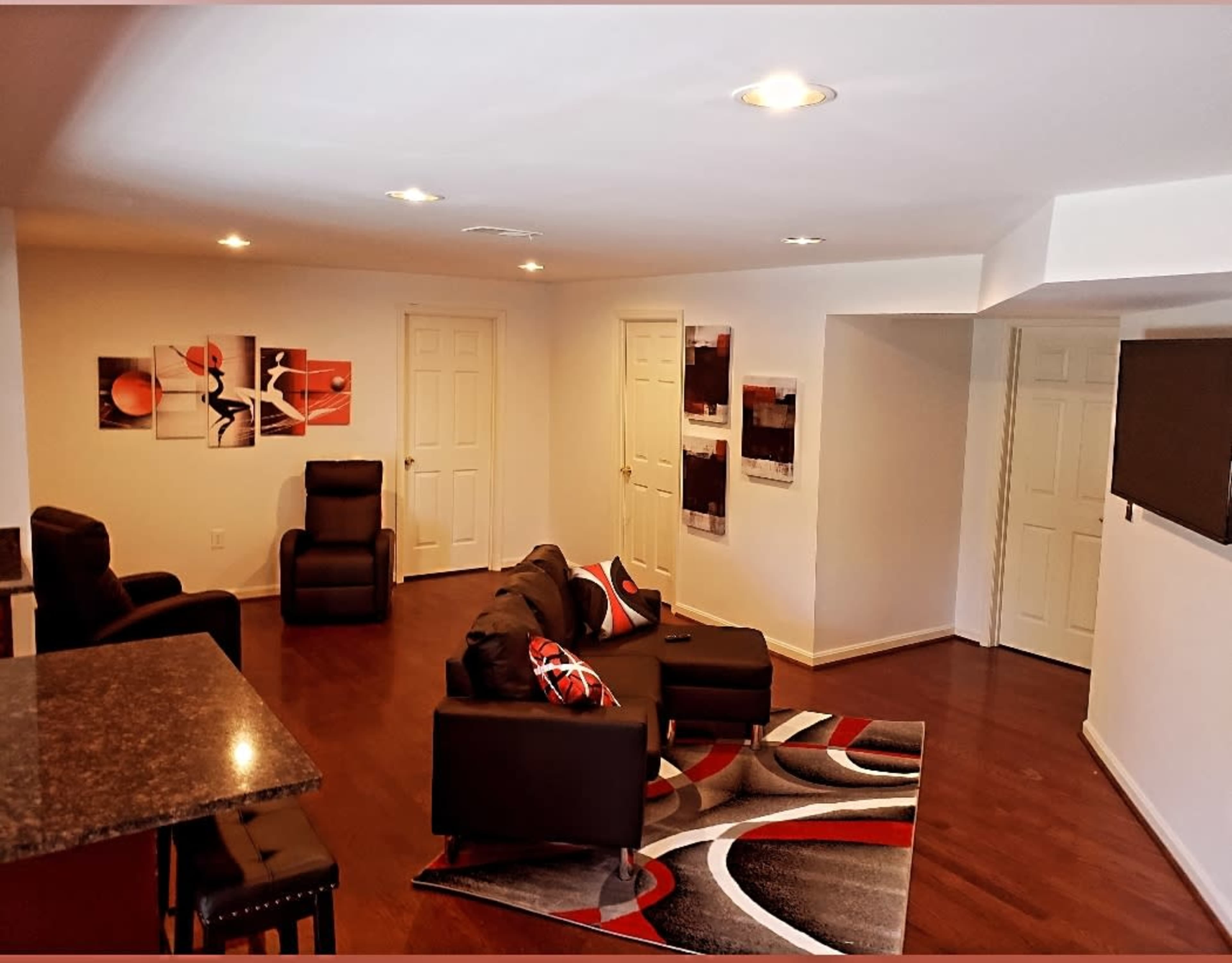 A modern living room with hardwood floors, a sectional sofa, a patterned rug, and two reclining chairs, along with wall art and a television mounted on the wall.