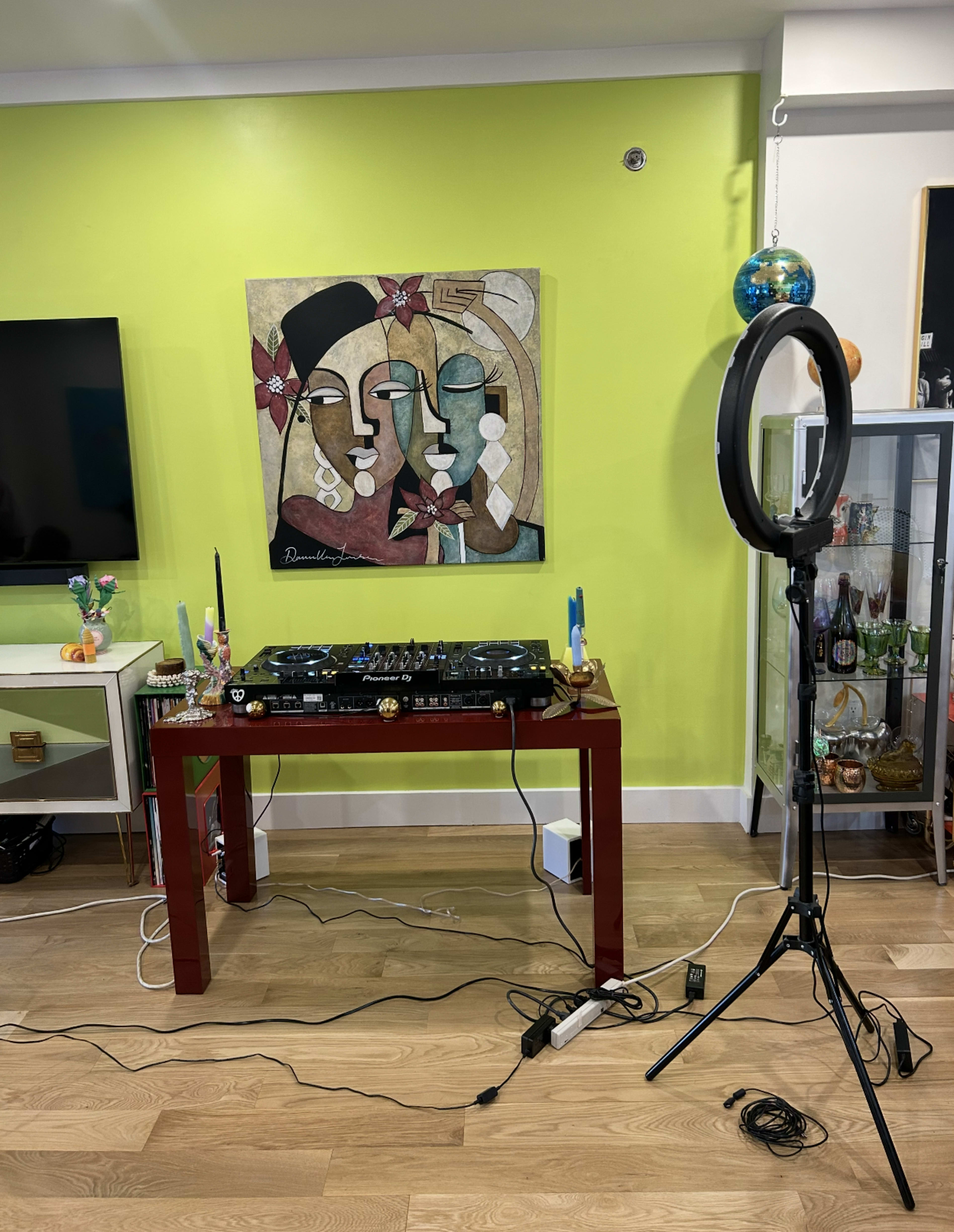 A DJ setup with turntables is placed on a red table beneath an abstract painting on a bright green wall, alongside a ring light and various decorative items.