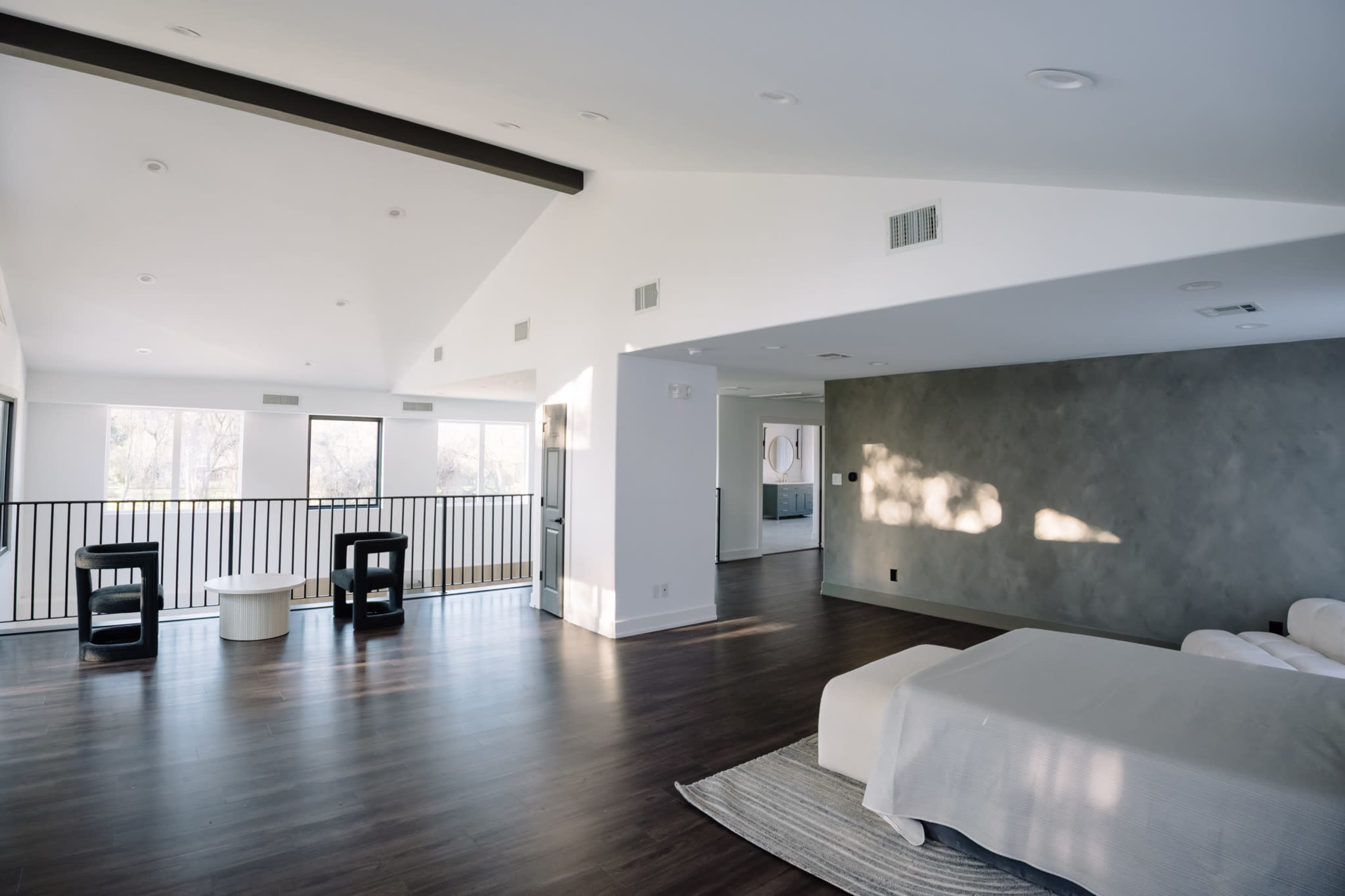 The image shows a spacious, modern bedroom with wood flooring, large windows, and minimal furnishings, including a bed, a sofa, and two black side tables.