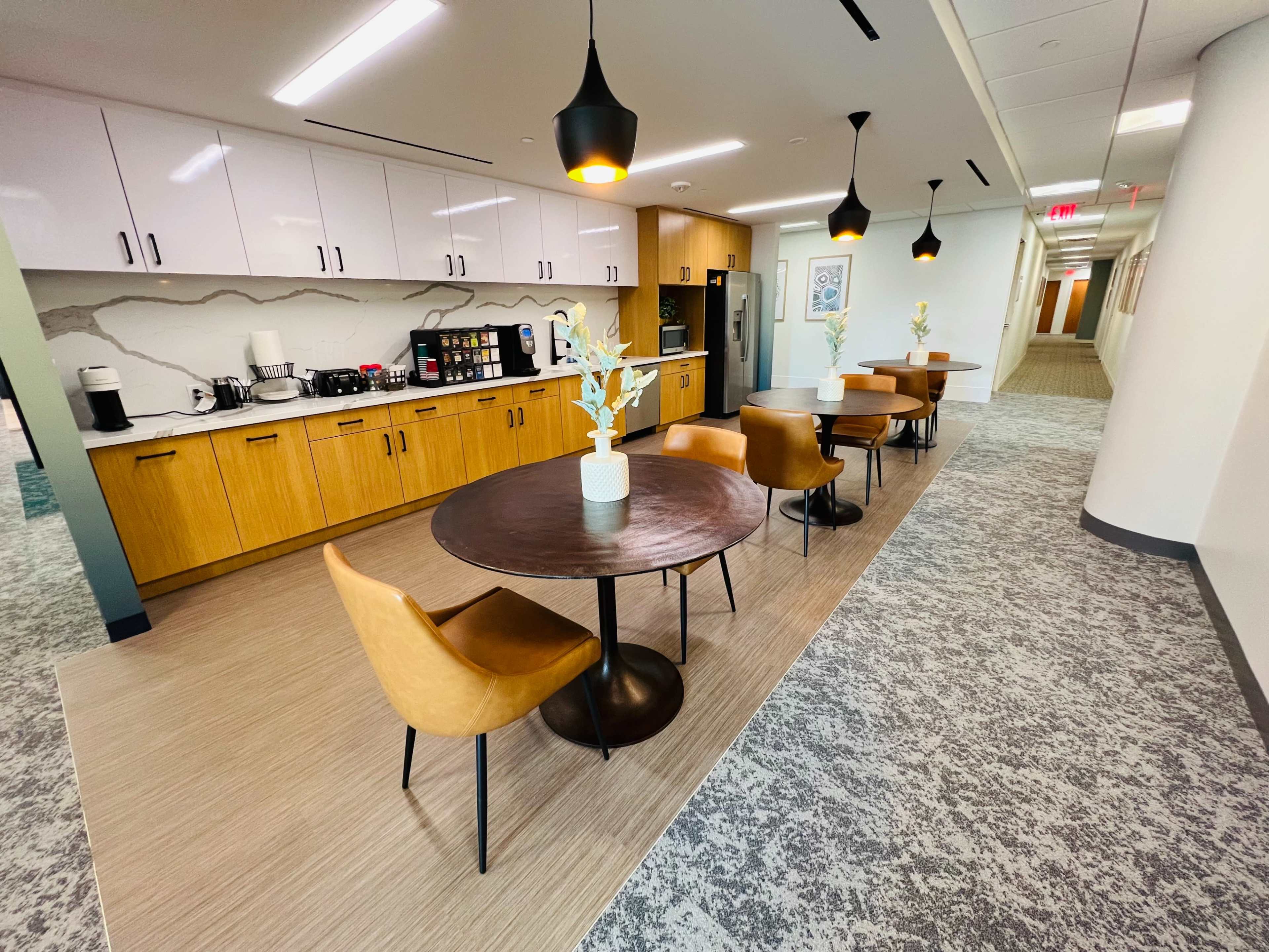 A modern office kitchen area featuring wooden cabinetry, a coffee station, and a dining space with a round table and chairs.