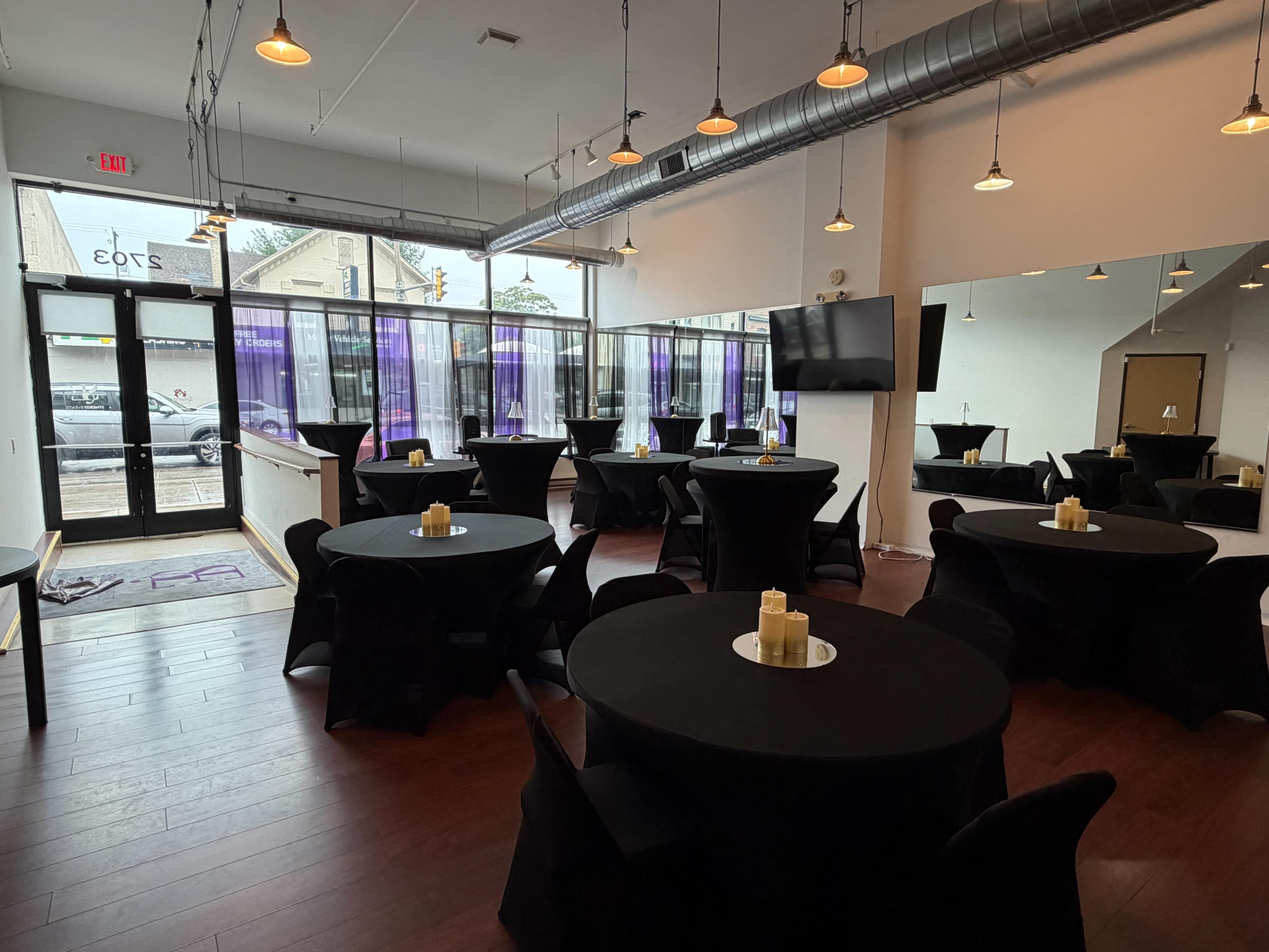 The image shows a modern event space with several round tables covered in black tablecloths, each adorned with a candle centerpiece, and large windows letting in natural light.