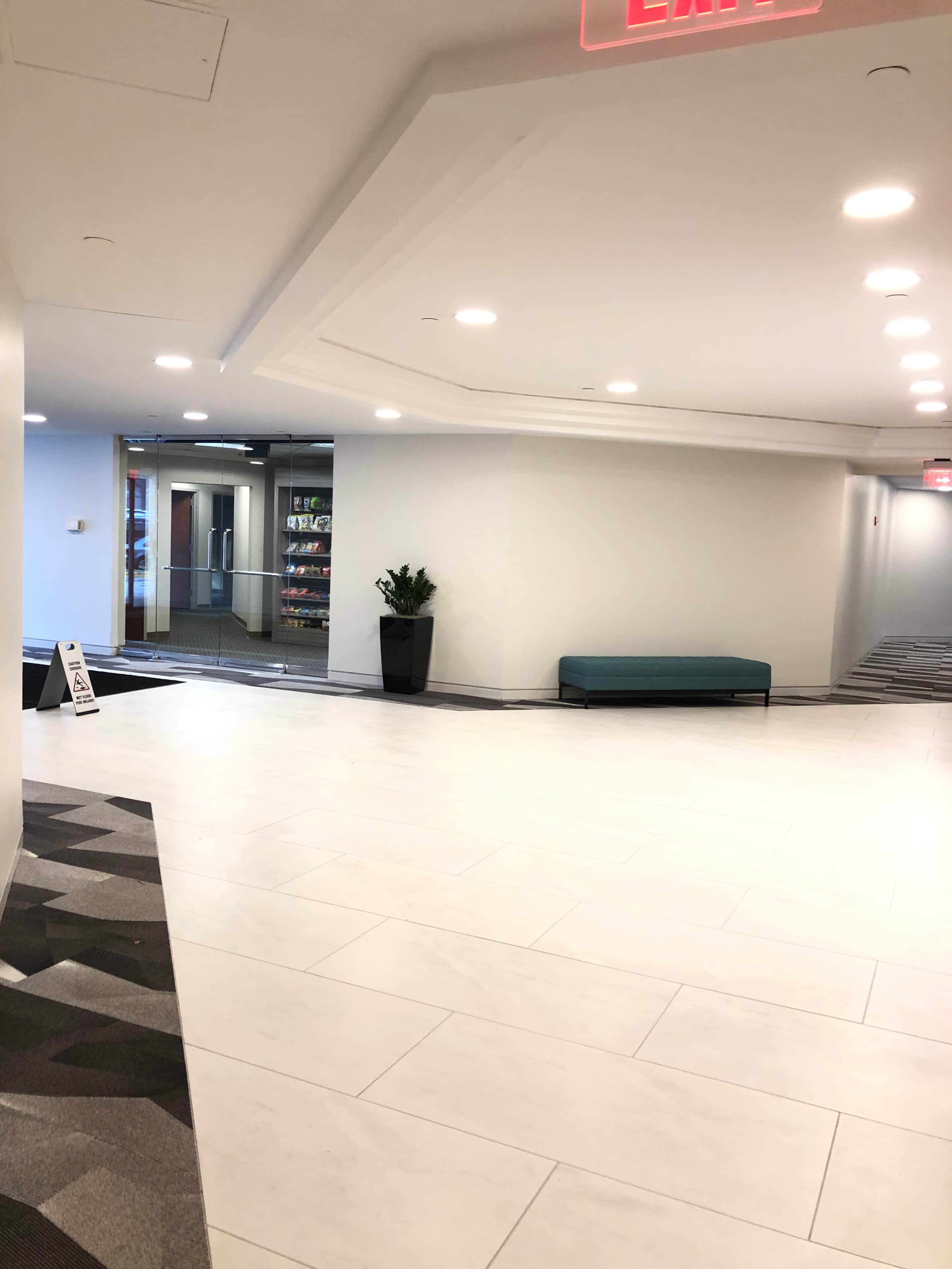 A modern hallway with white tiled flooring, a small plant in a pot, a teal bench, and a glass door leading to another room.