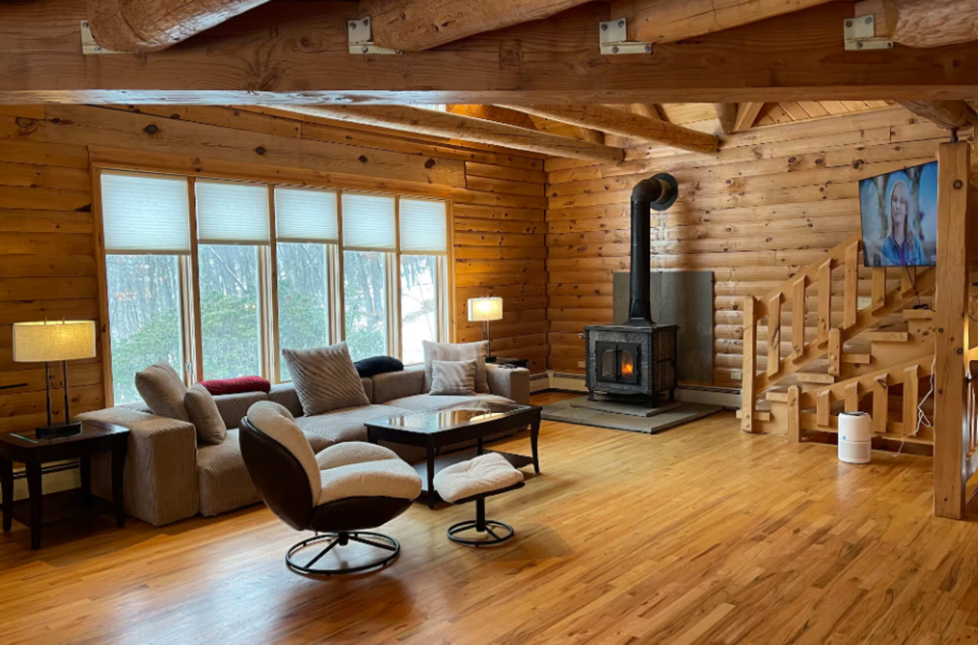 The image shows a cozy log cabin living room with a fireplace, large windows, and a staircase in the background.