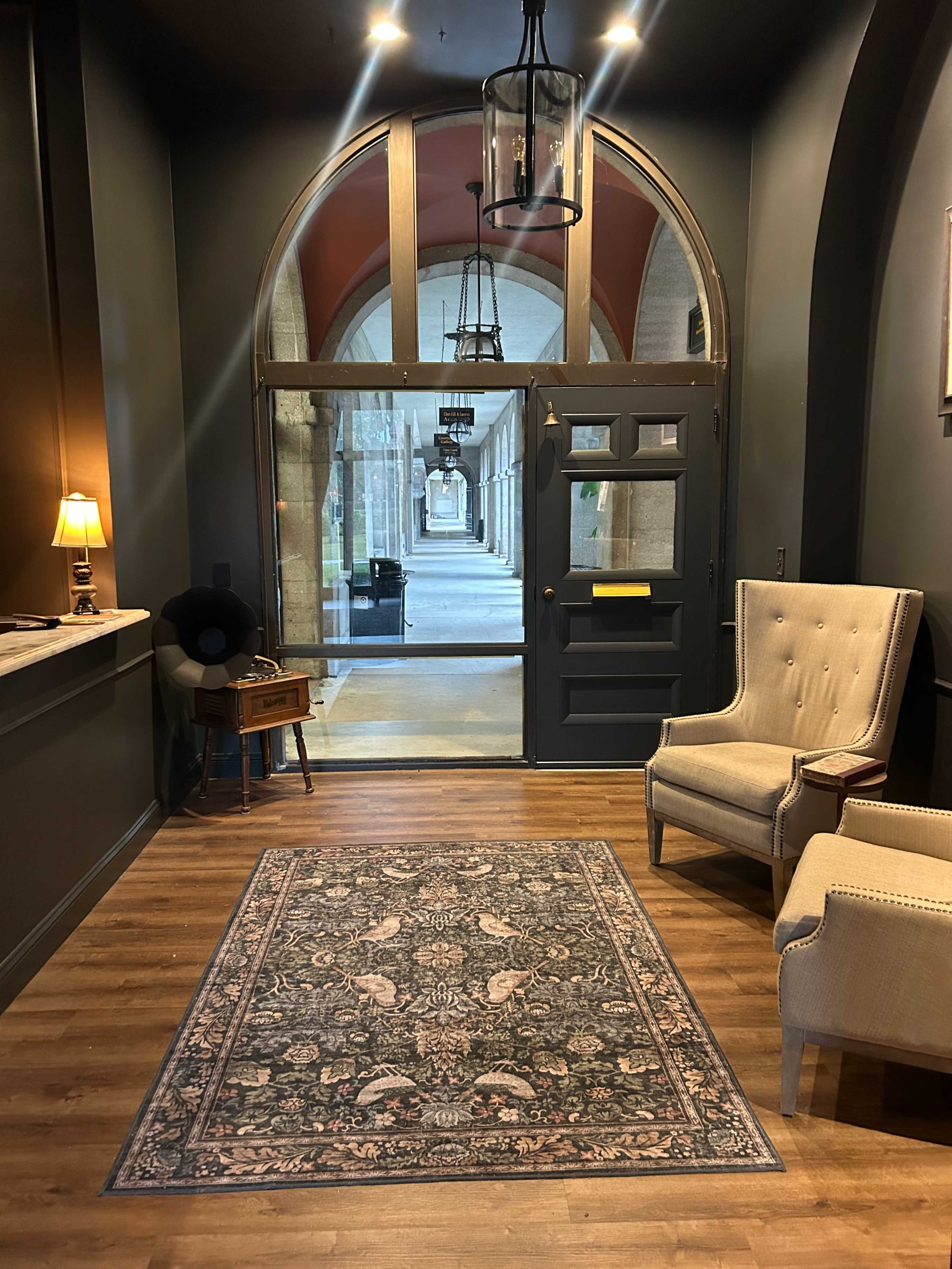 The image shows a cozy interior lobby with two armchairs, a patterned rug, a desk, and a large arched window that frames a view of a corridor.