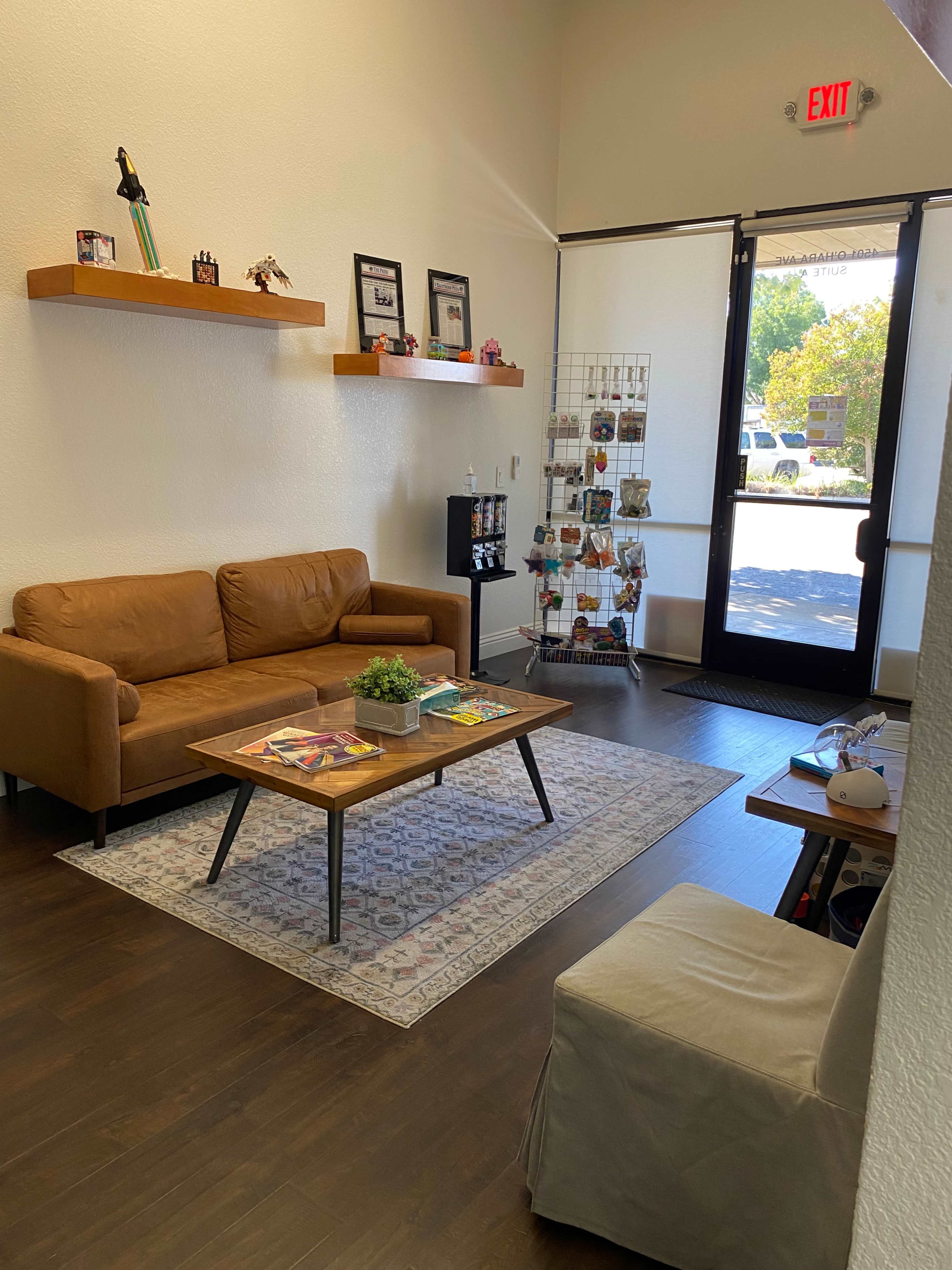 A waiting area features two brown couches, a coffee table with magazines, a decorative rug, and a display rack of postcards by the entrance.