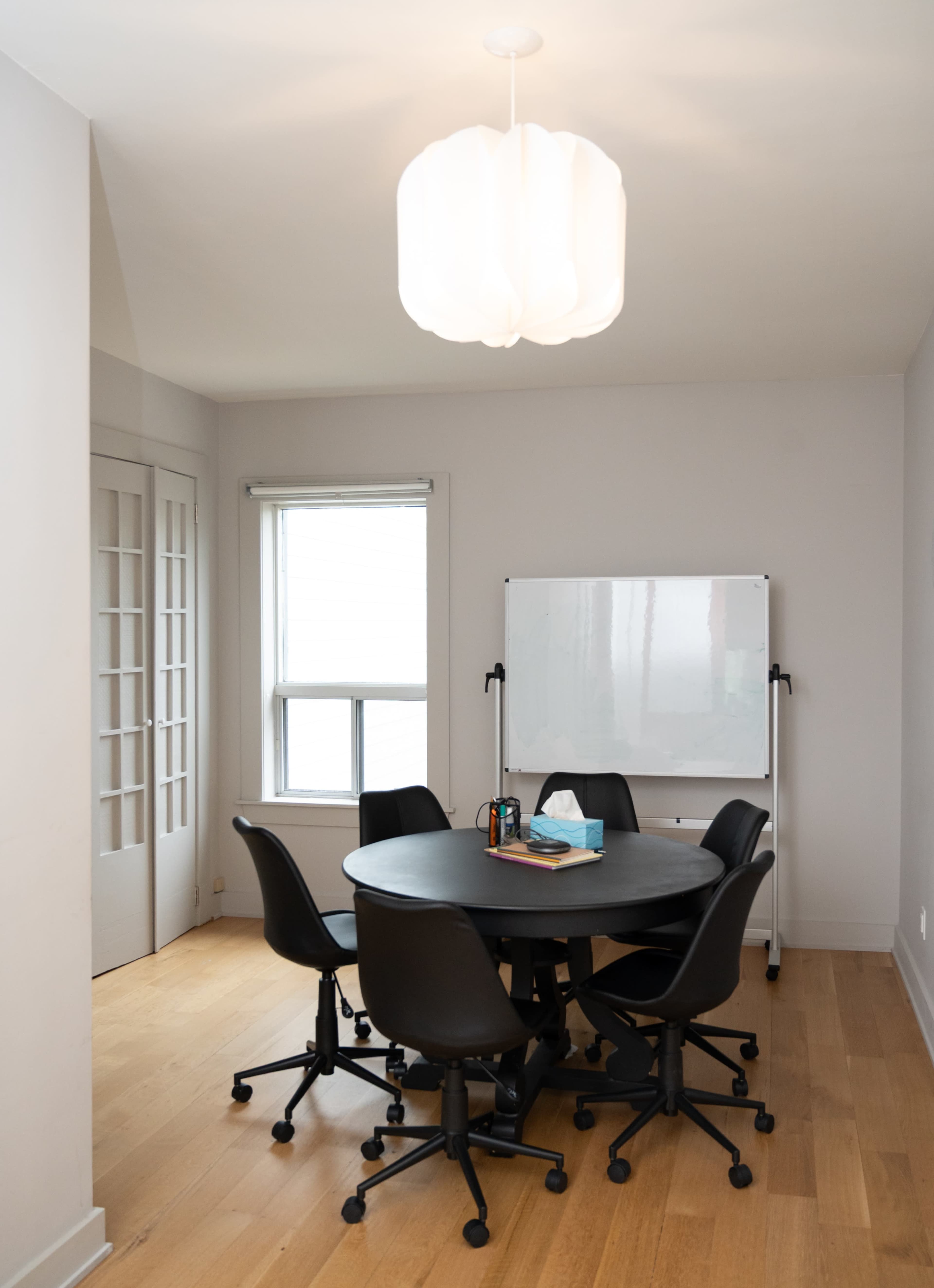 The image shows a modern meeting room with a round table surrounded by five black chairs, a whiteboard on a stand, and a hanging light fixture.