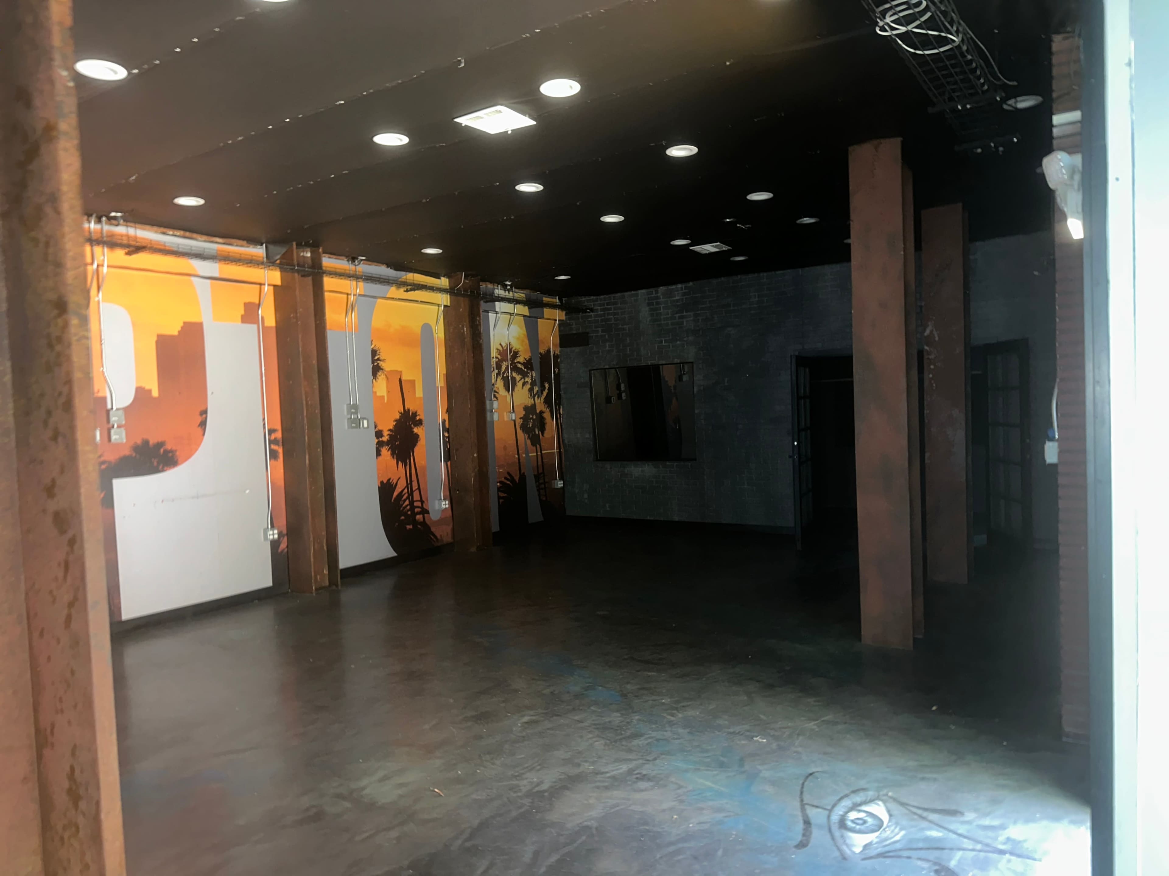 The image shows an industrial-style indoor space with dark flooring and exposed pillars, featuring a vibrant sunset mural on one wall.