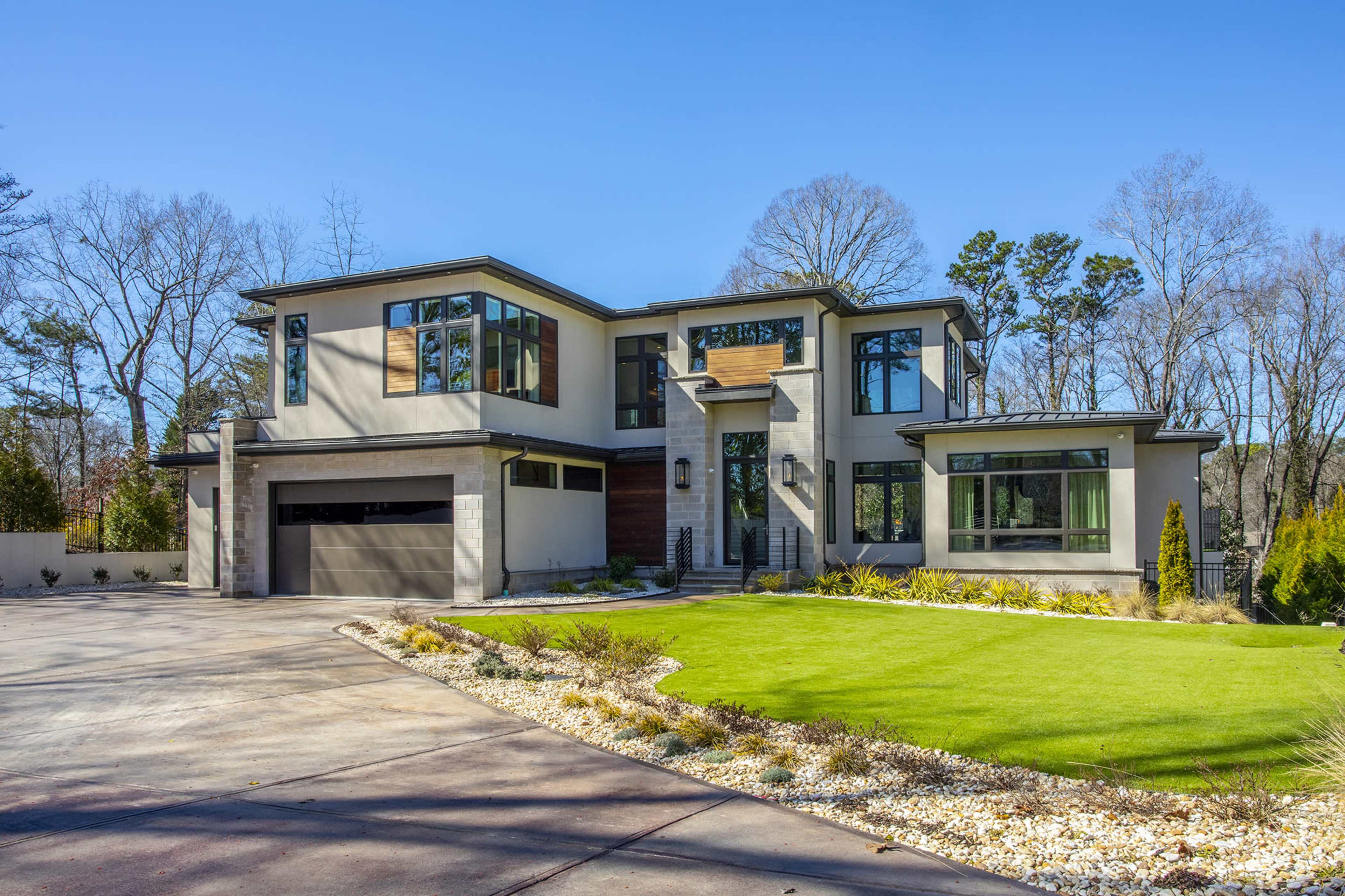 A modern two-story house features large windows, a stone and wood facade, and a well-manicured lawn with landscaped gardens.