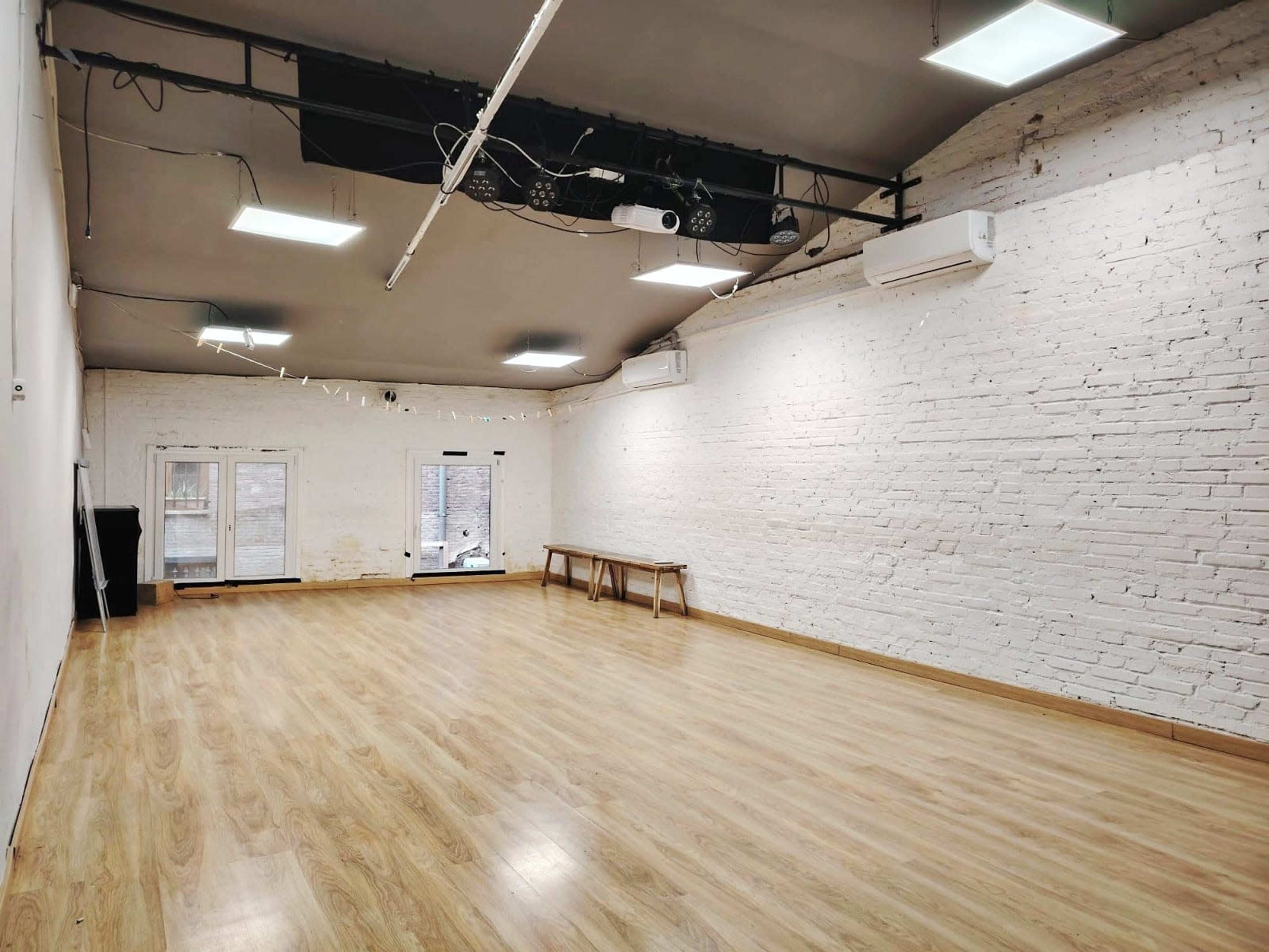 The image shows a spacious, empty room with white brick walls, a wooden floor, and large windows at one end.