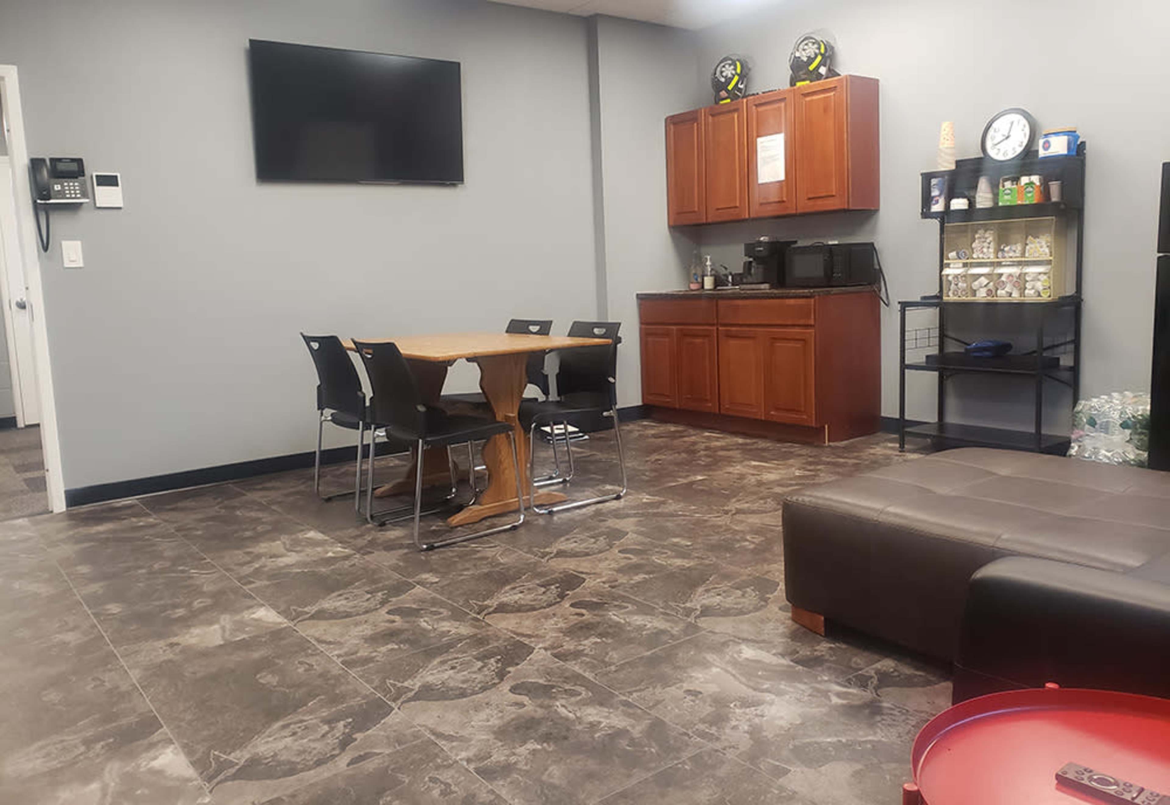 A break room with a wooden dining table and chairs, a kitchenette with cabinets, a microwave, and a television mounted on the wall.