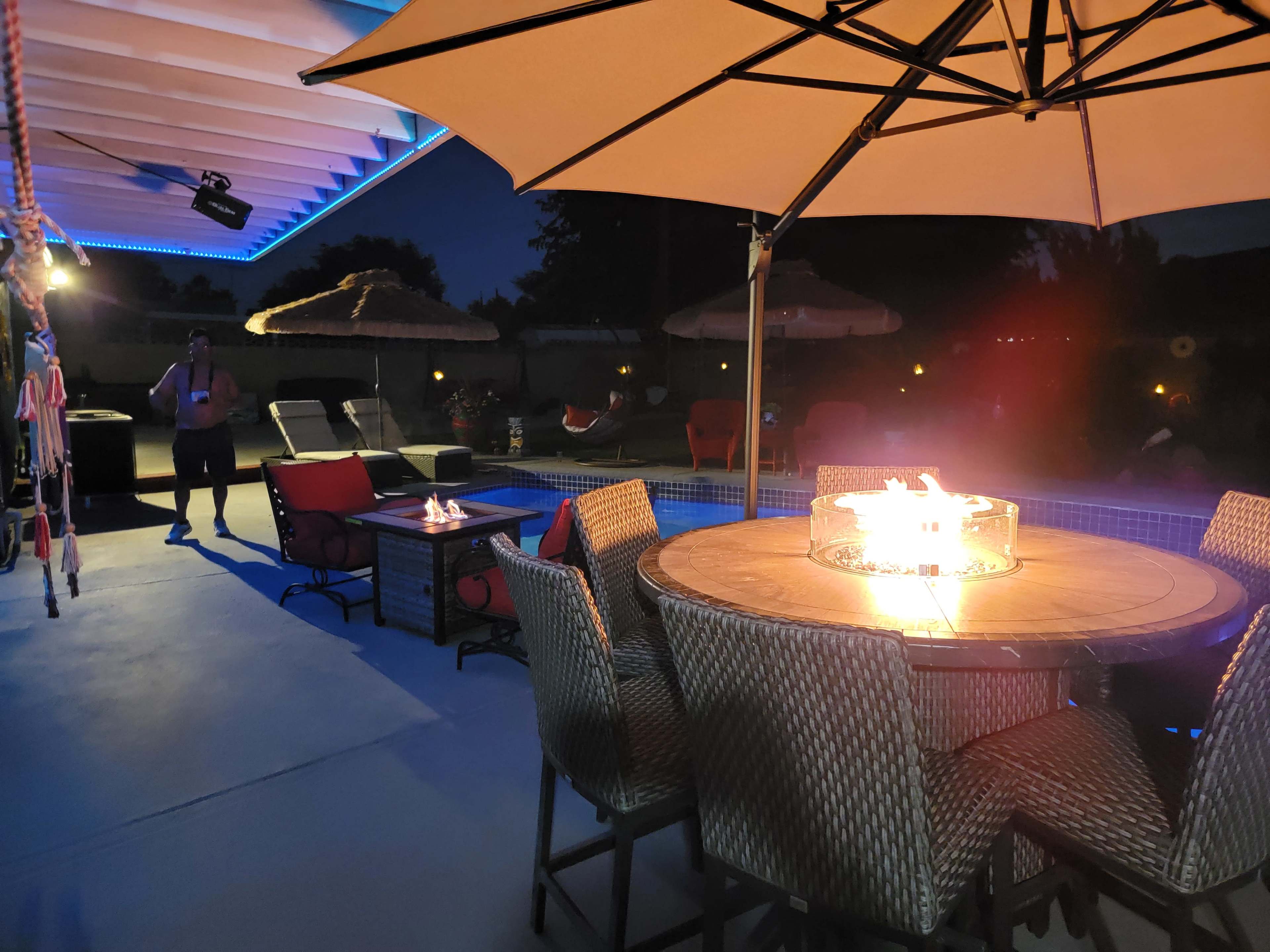 A patio area features a circular fire pit at a table surrounded by chairs, with lounge chairs and umbrellas in the background under evening lighting.