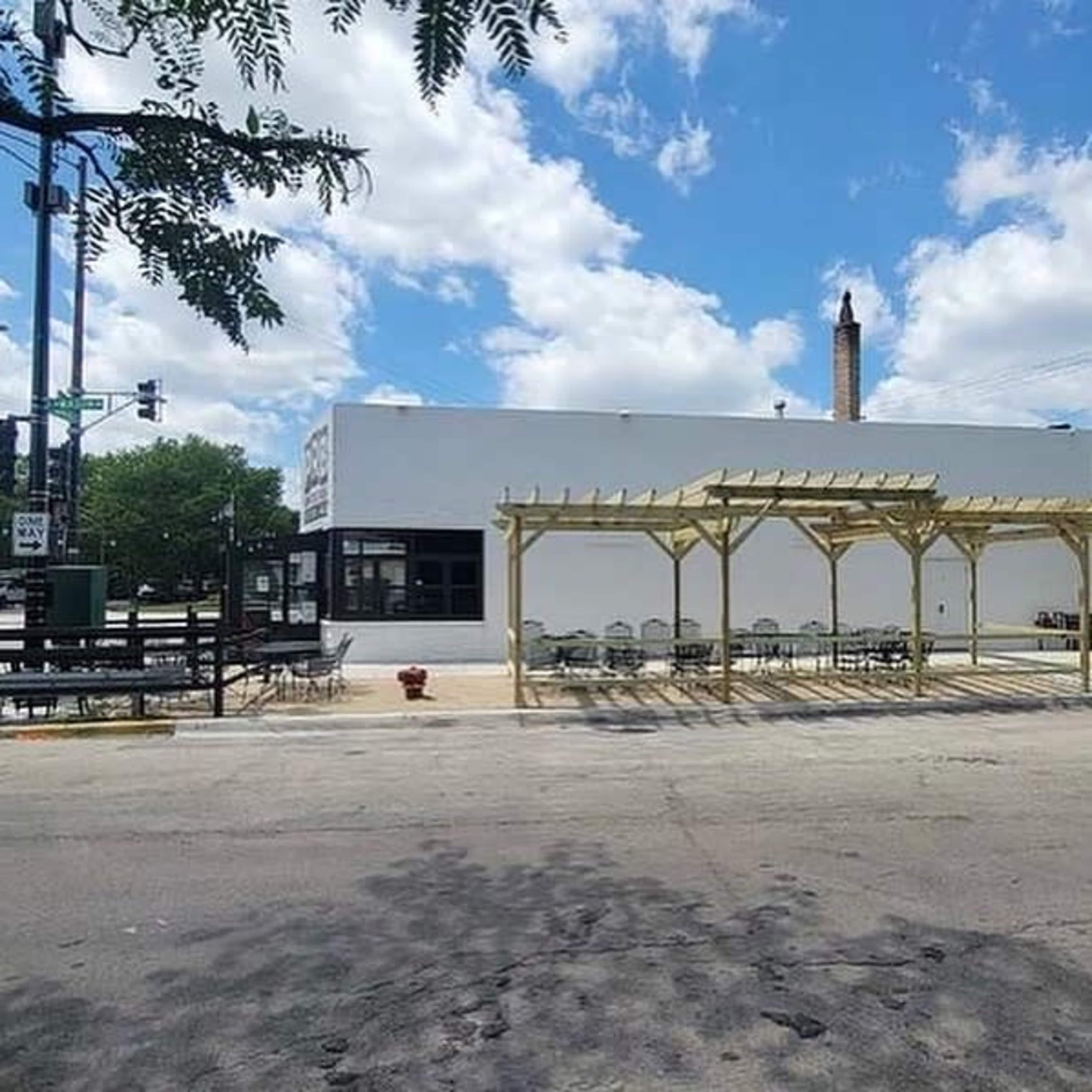 A modern building with a white exterior and a wooden pergola is located on a street corner, with outdoor seating arranged beneath it.