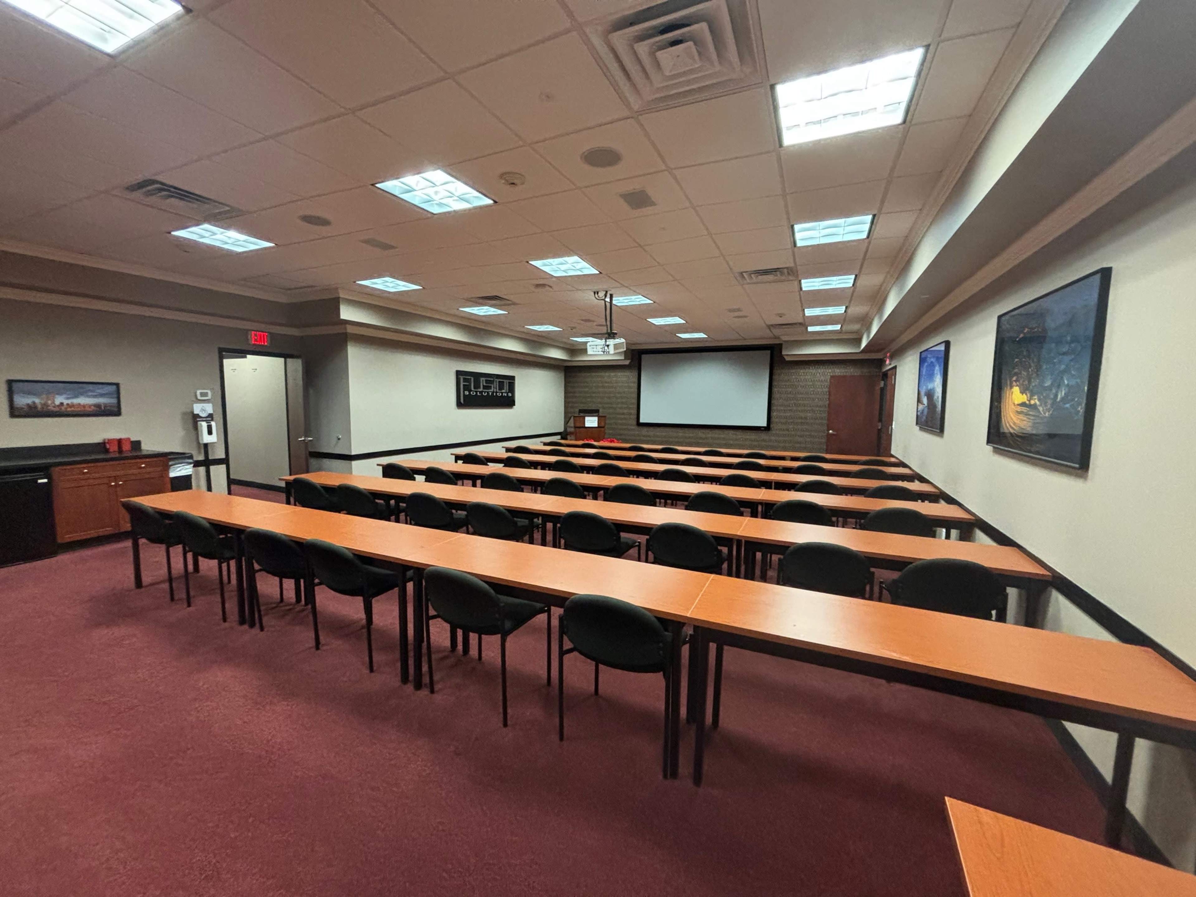 A conference room featuring multiple rows of tables and chairs, with a projector screen and wall-mounted artwork.