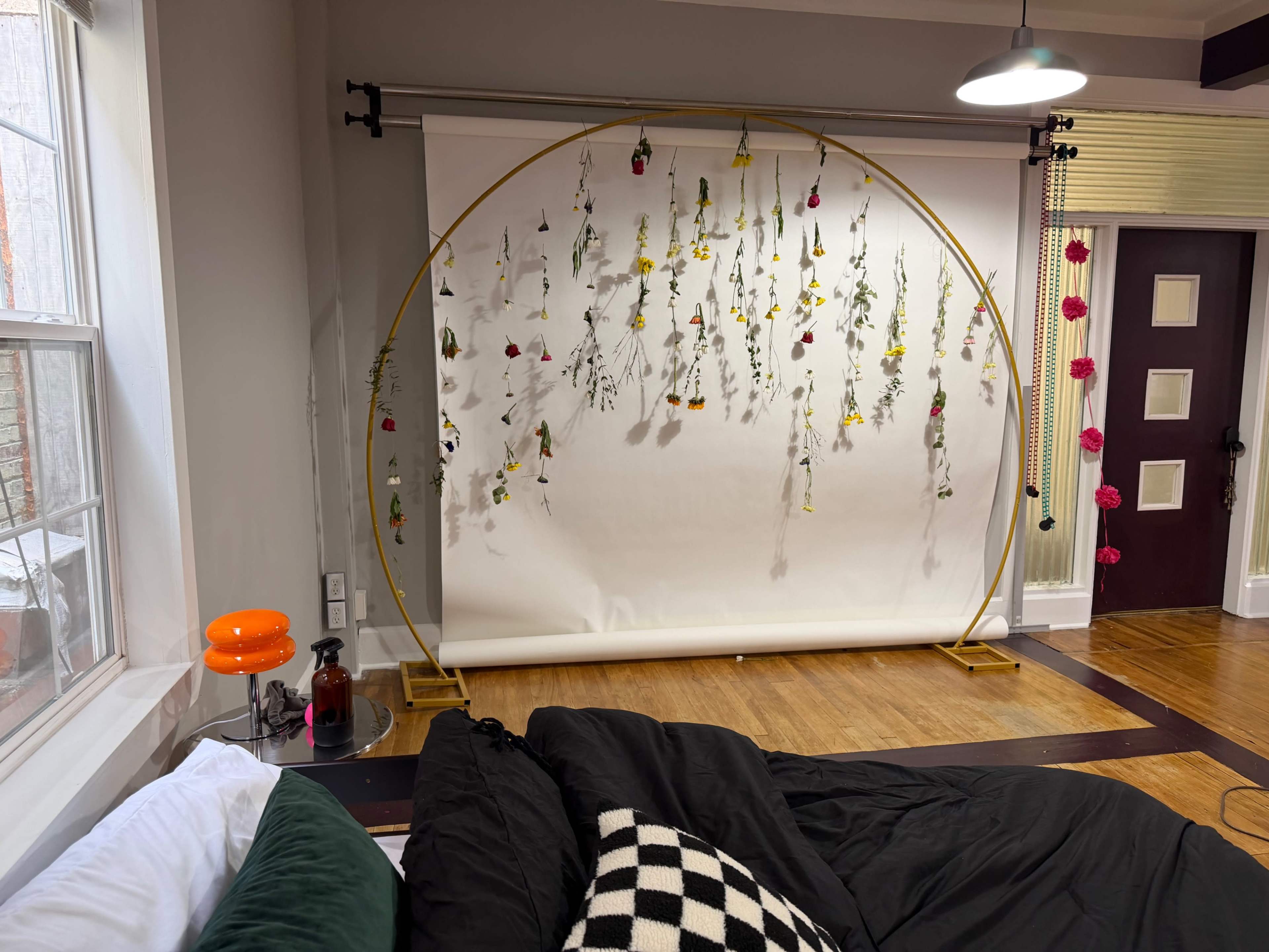 The image shows a room with a black bed, a vintage orange table lamp, and a white backdrop adorned with hanging flowers, set against a wooden floor and a wall with a door.