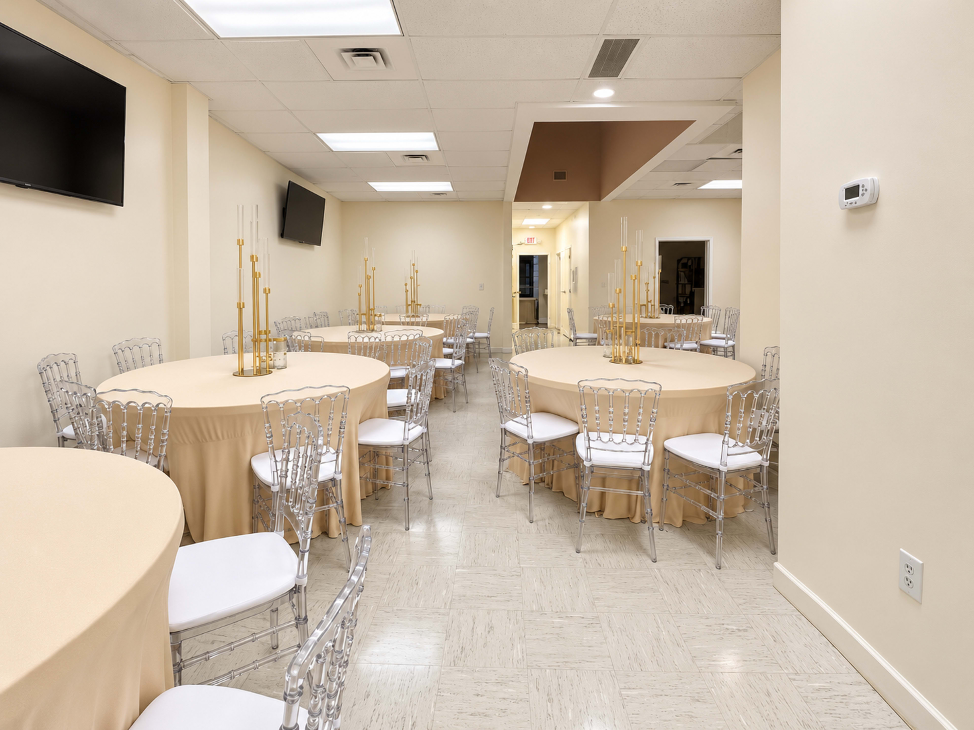 The image features a spacious event room with several round tables set with beige tablecloths and clear acrylic chairs, illuminated by overhead lighting.
