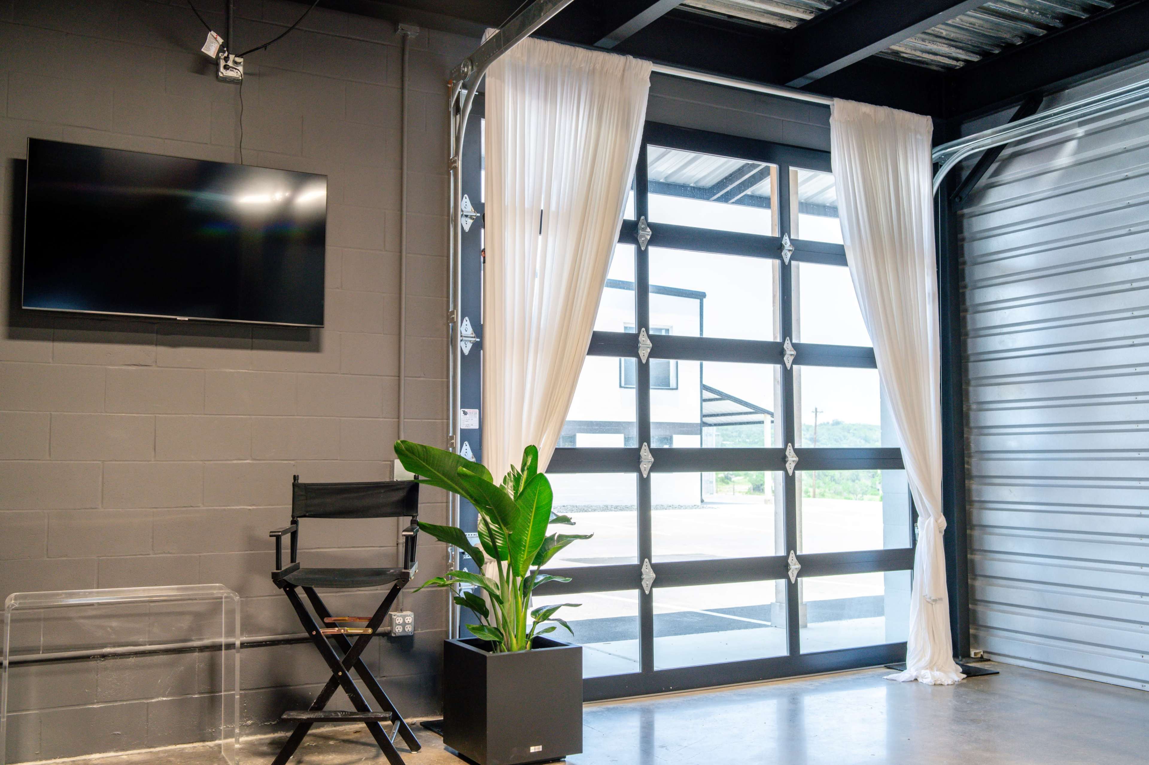 A modern interior space with a flat-screen TV mounted on a wall, a large glass door covered with sheer curtains, a folding chair, and a potted plant.