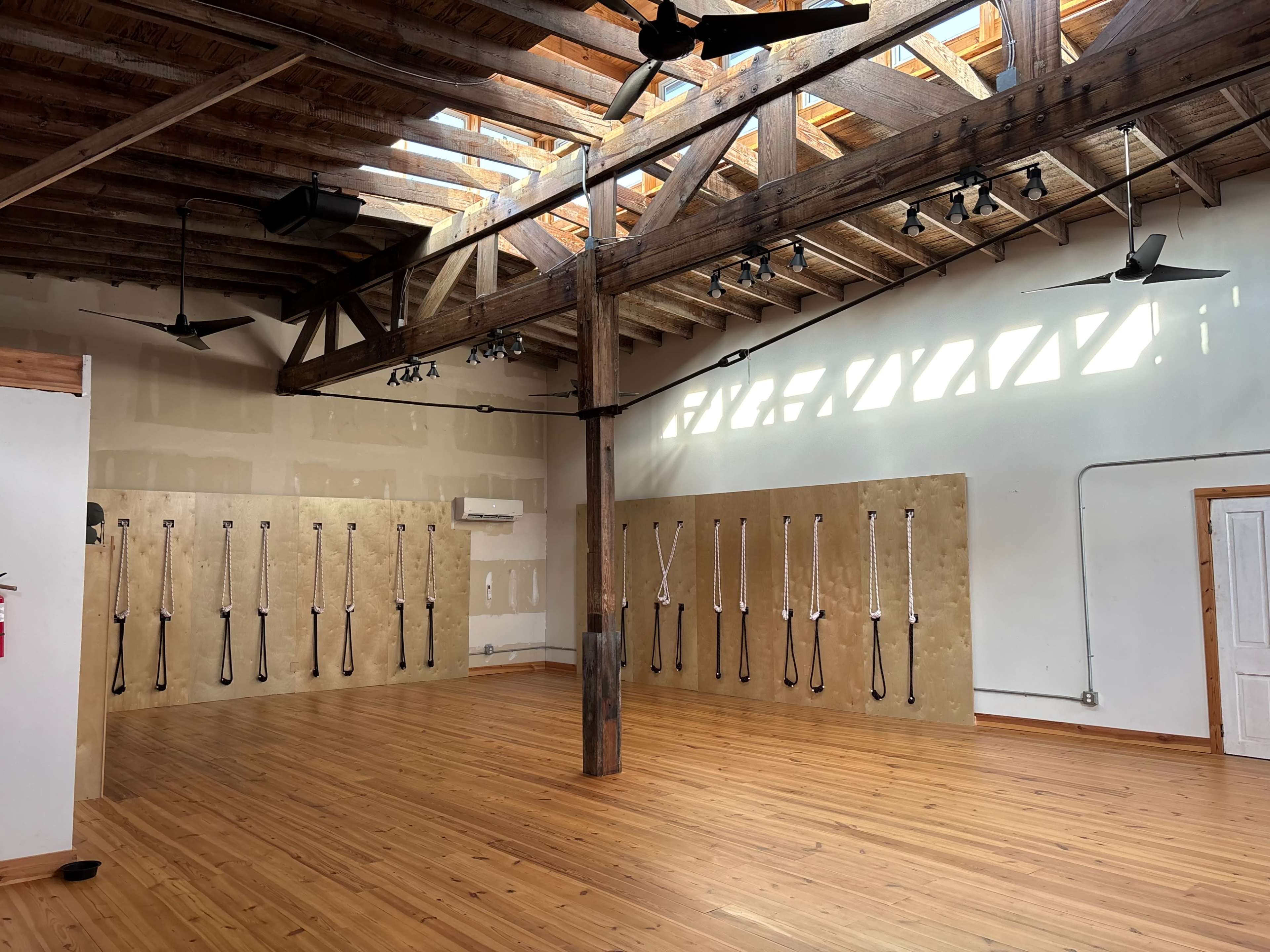 The image shows a spacious room with wooden floors and high ceilings, featuring wooden beams and multiple fans, along with a wall displaying suspended straps for fitness activities.