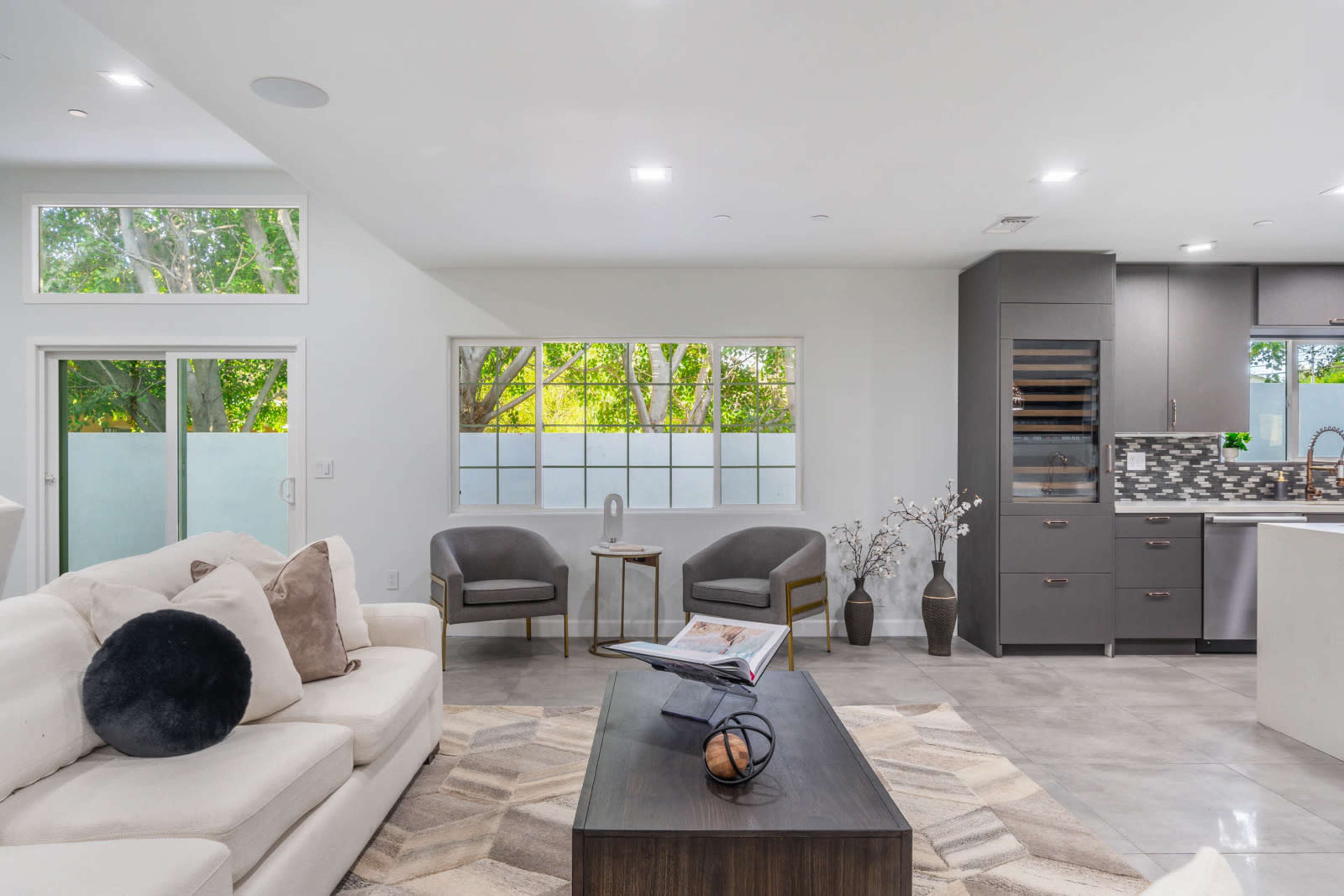 The image shows a modern living area featuring a light-colored sofa, two chairs near a window, a coffee table with a book, and a kitchen area in the background.
