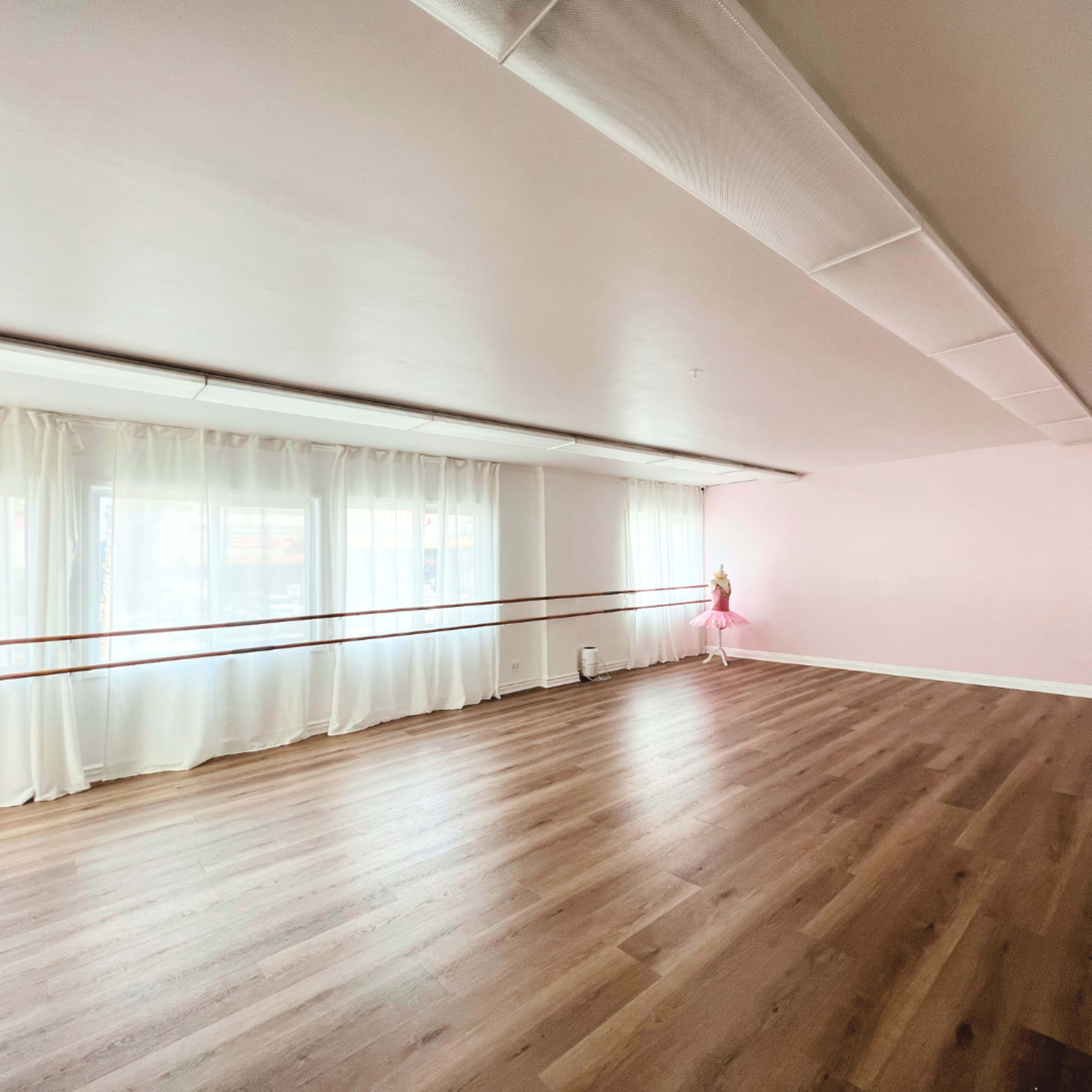 A spacious dance studio features wooden flooring, large windows with white curtains, and a pink ballet tutu displayed in the corner.