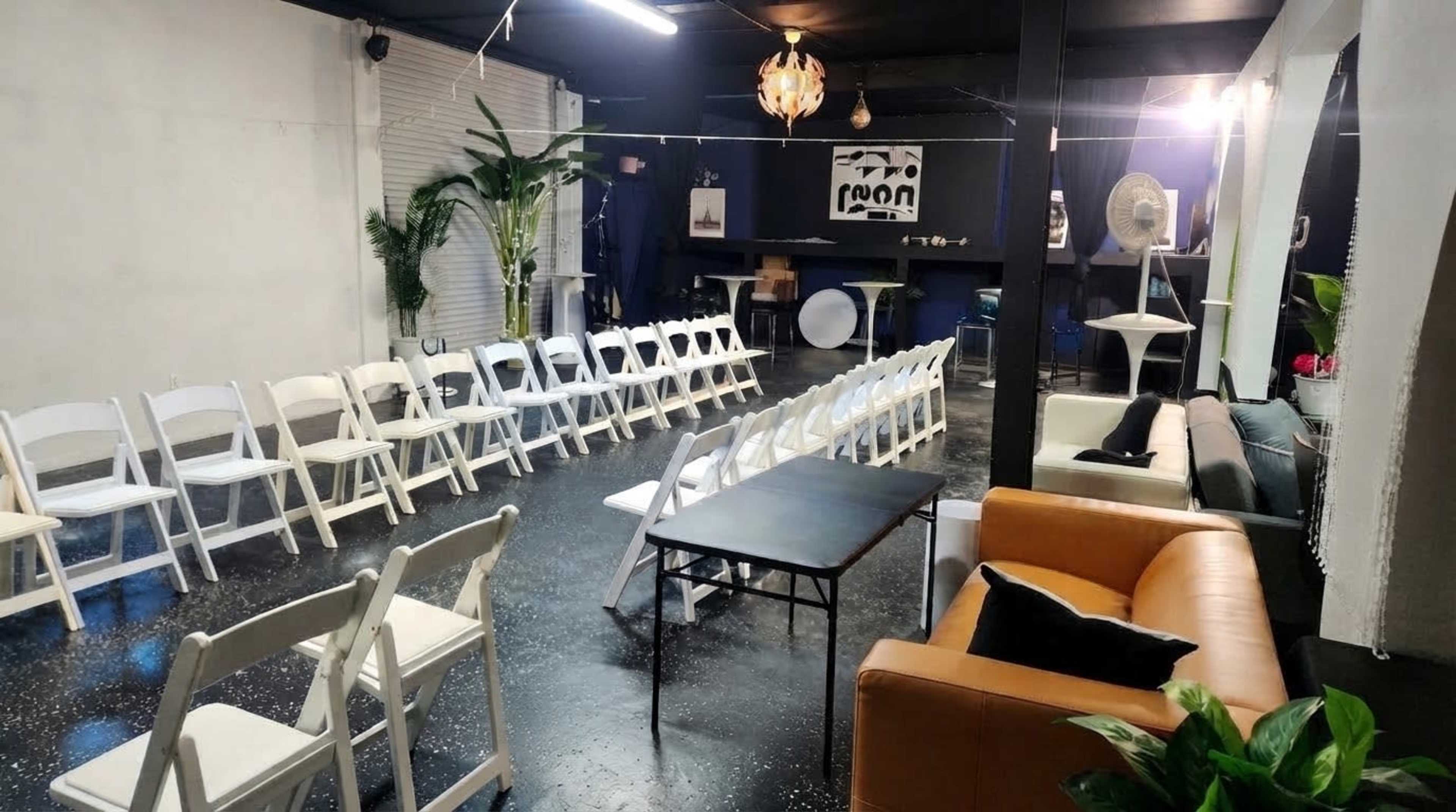 The image shows a spacious indoor venue with rows of white chairs facing a central table, surrounded by dark walls and decorative plants.
