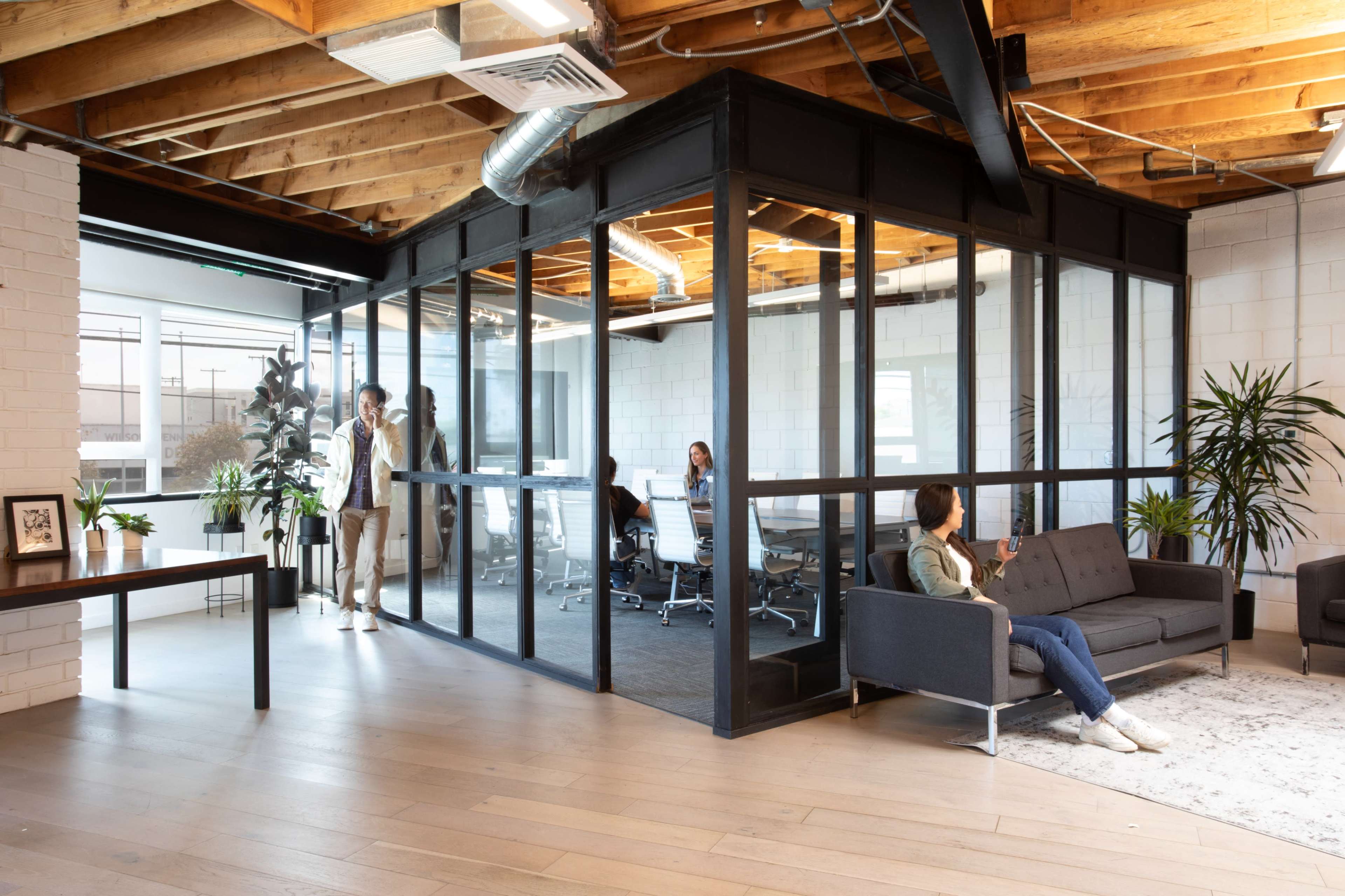 The image shows a modern office space featuring a glass-walled conference room and comfortable seating areas with people engaged in work.