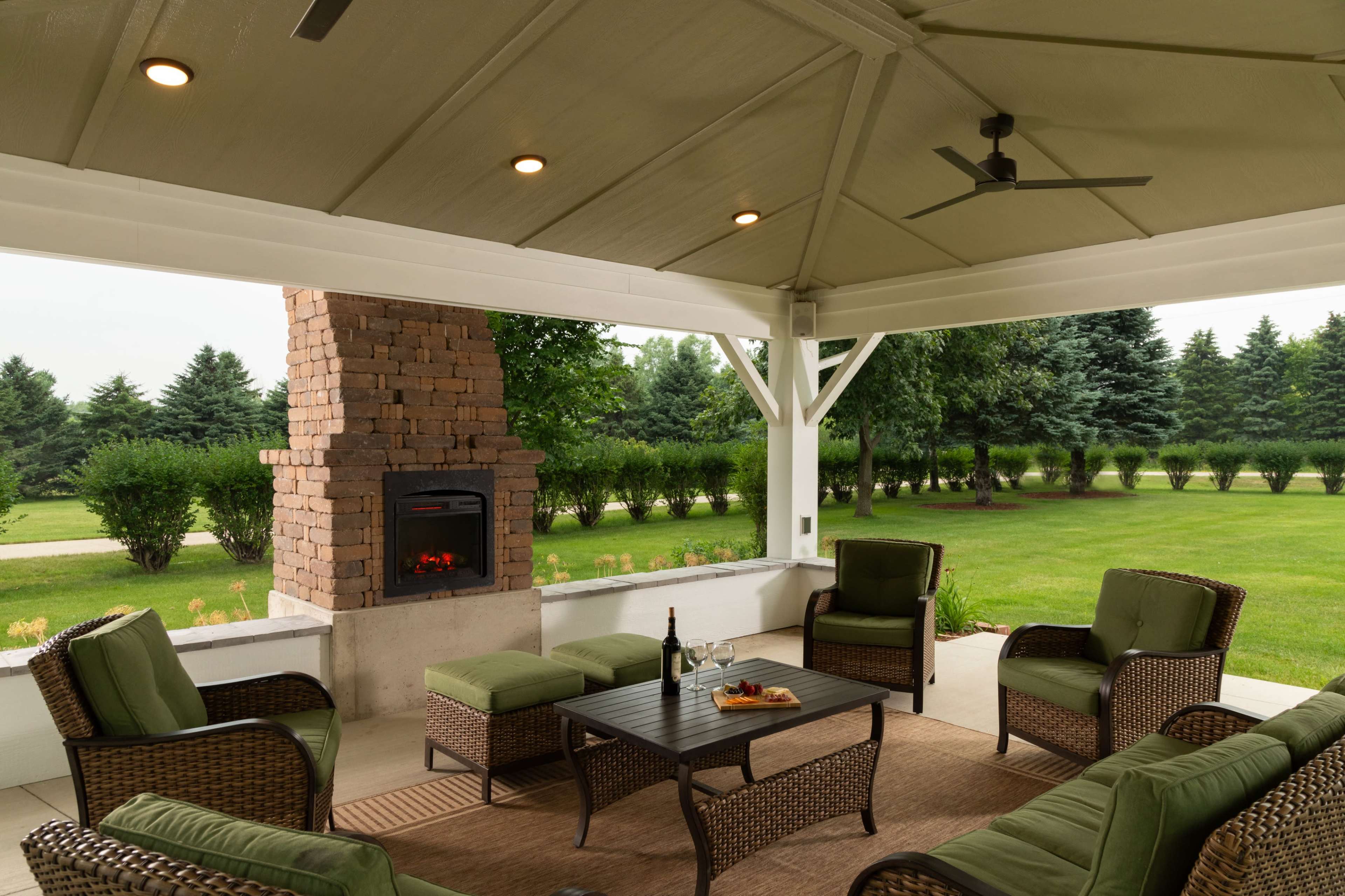 The image shows a covered patio with comfortable seating around a table, featuring a stone fireplace and a landscaped lawn in the background.