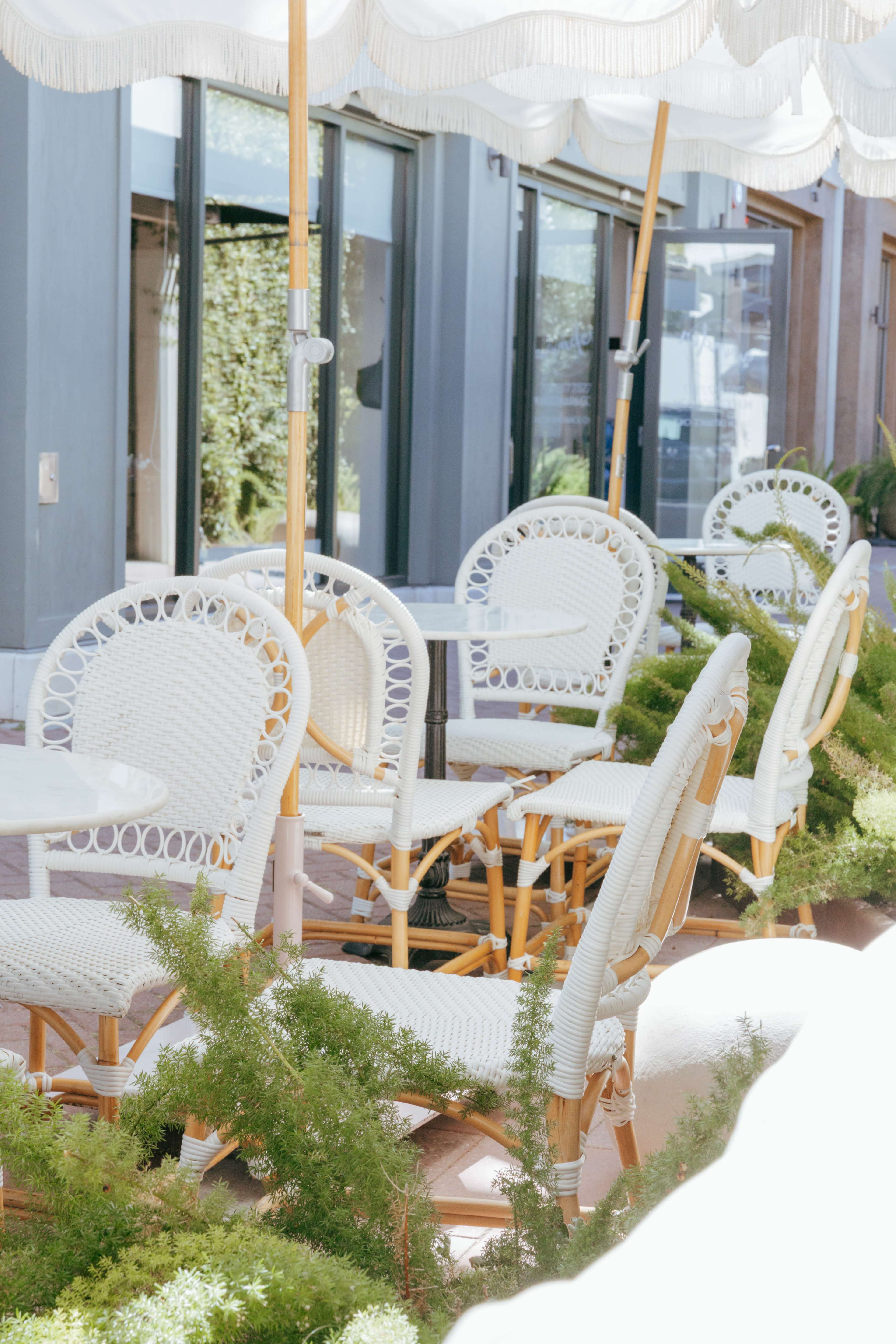 A set of white wicker chairs and a table are arranged under a large umbrella, with greenery surrounding them in an outdoor setting.