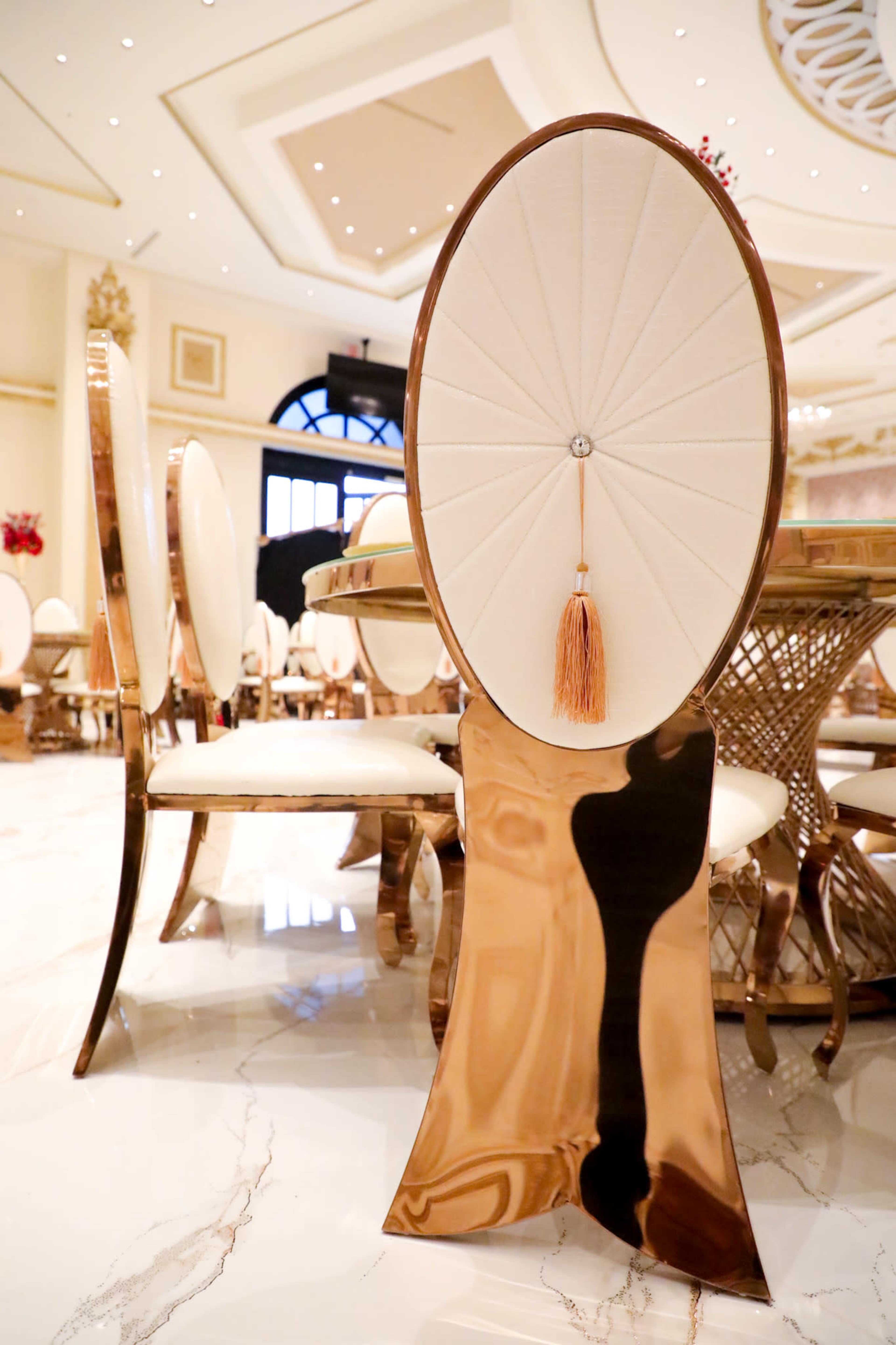 The image shows a decorative chair with a backrest featuring a fan-like design and a tassel, set in an elegant dining area with marble flooring.