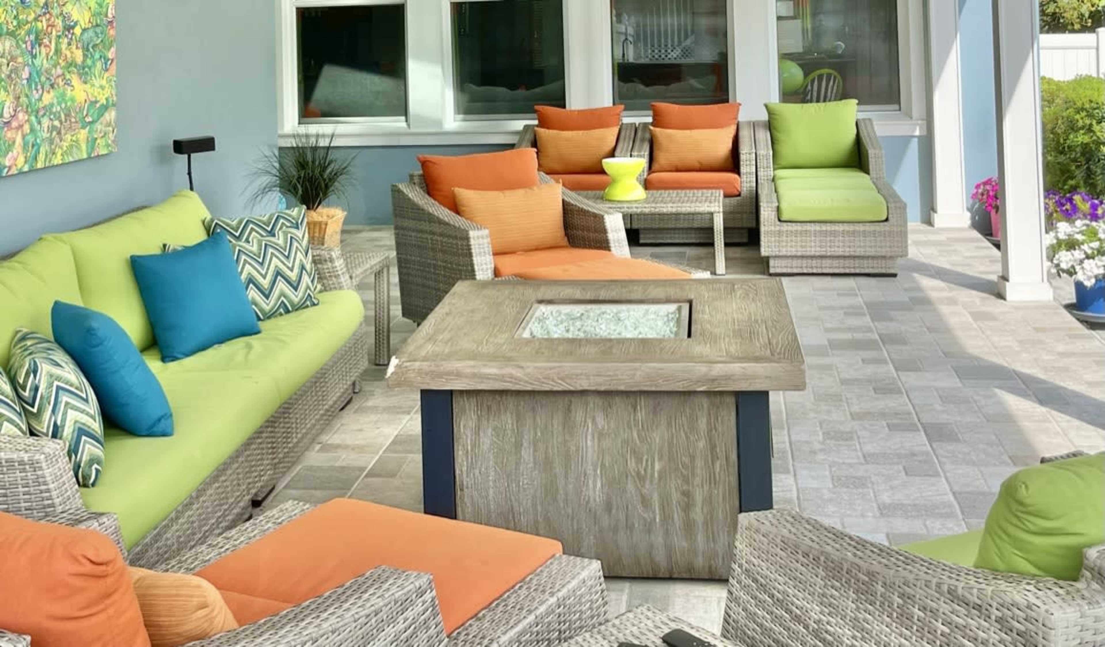 The image shows a cozy outdoor seating area with colorful cushions on wicker furniture surrounding a square table with a glass top.
