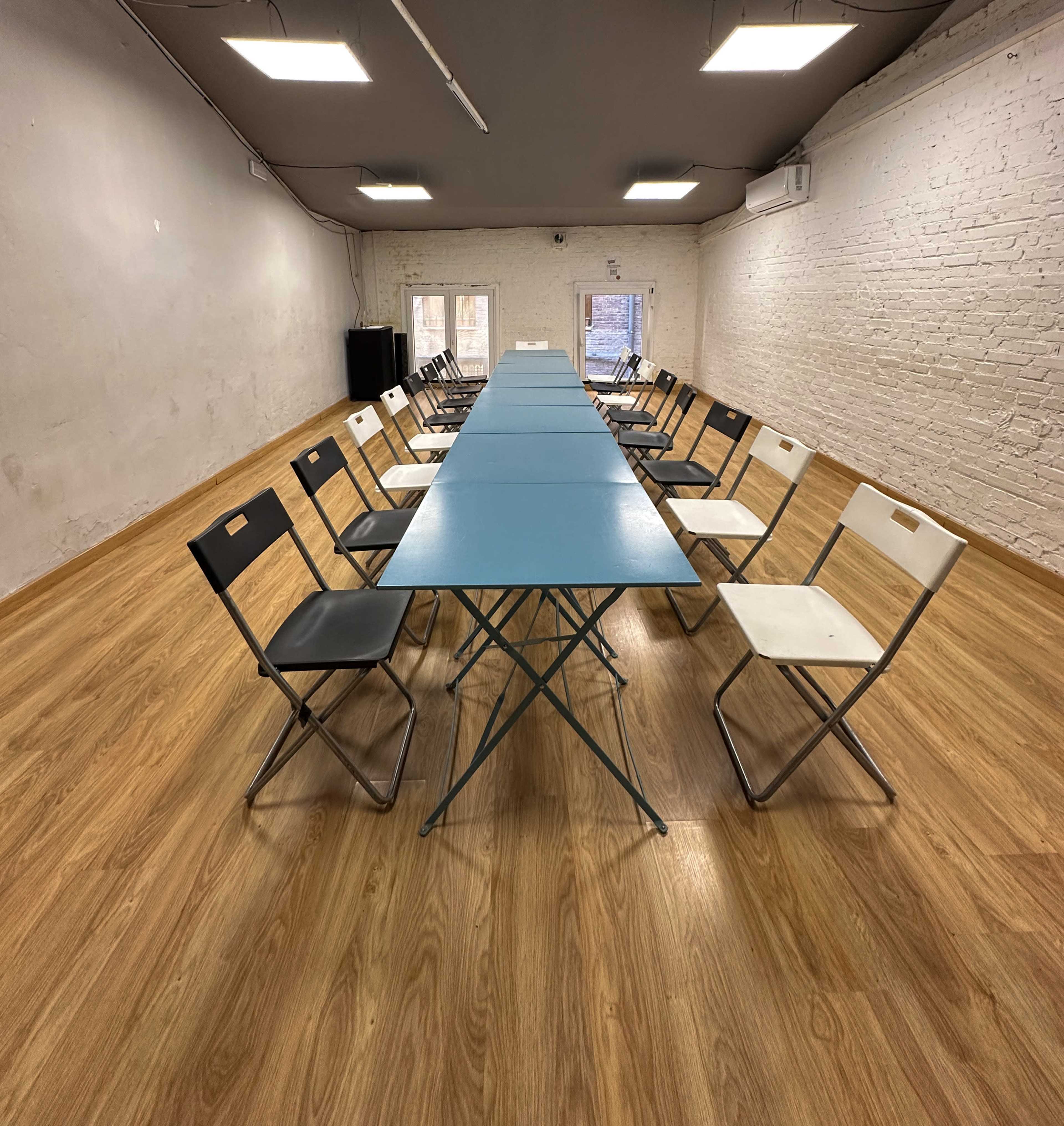 A long table with teal surface is surrounded by folding chairs in a minimalist interior space with wooden floors and white brick walls.