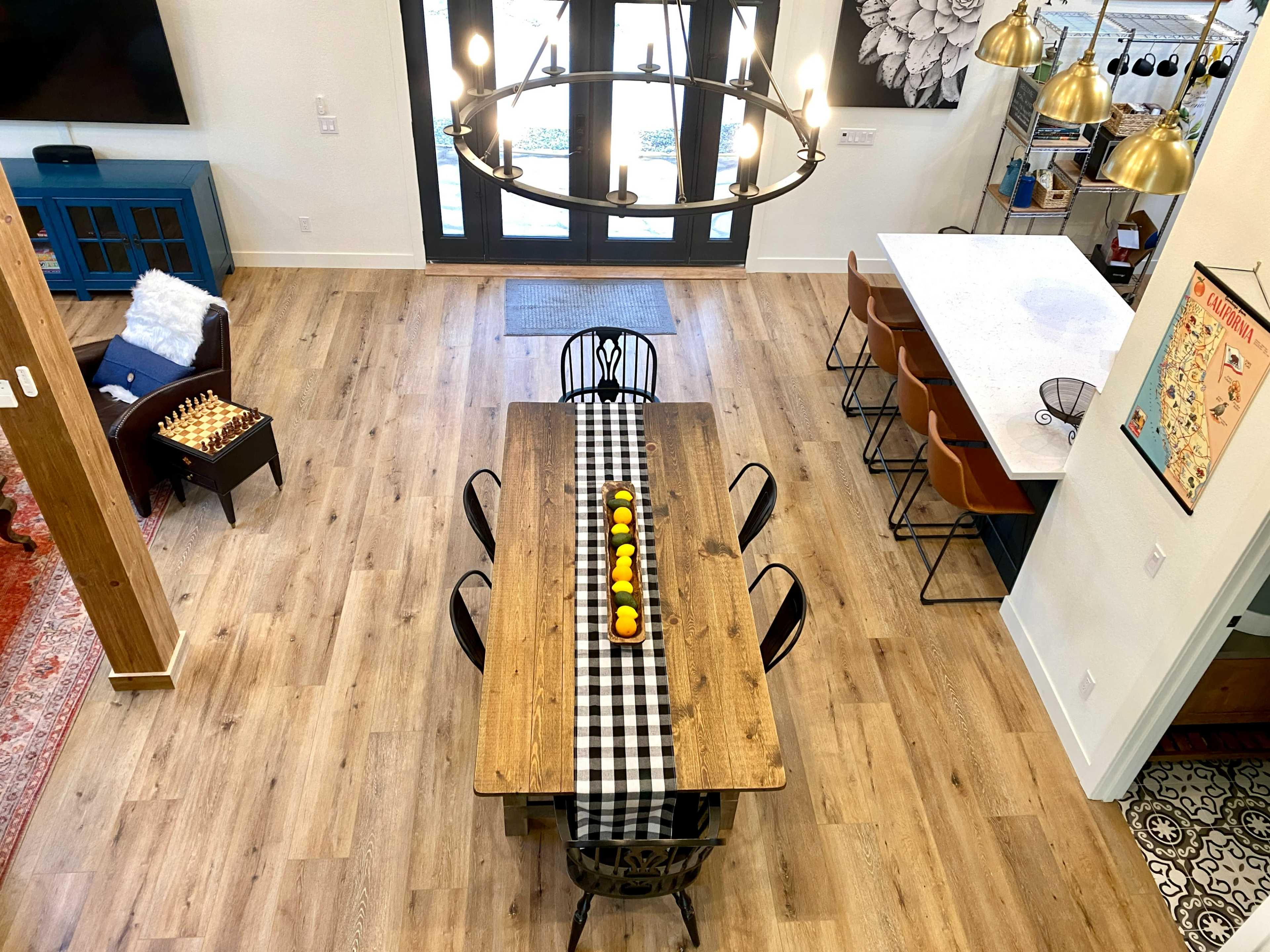 A spacious dining area features a long wooden table centered with a black and white checkered runner and decorative fruit, while modern lighting fixtures hang above and a kitchen bar is visible in the background.