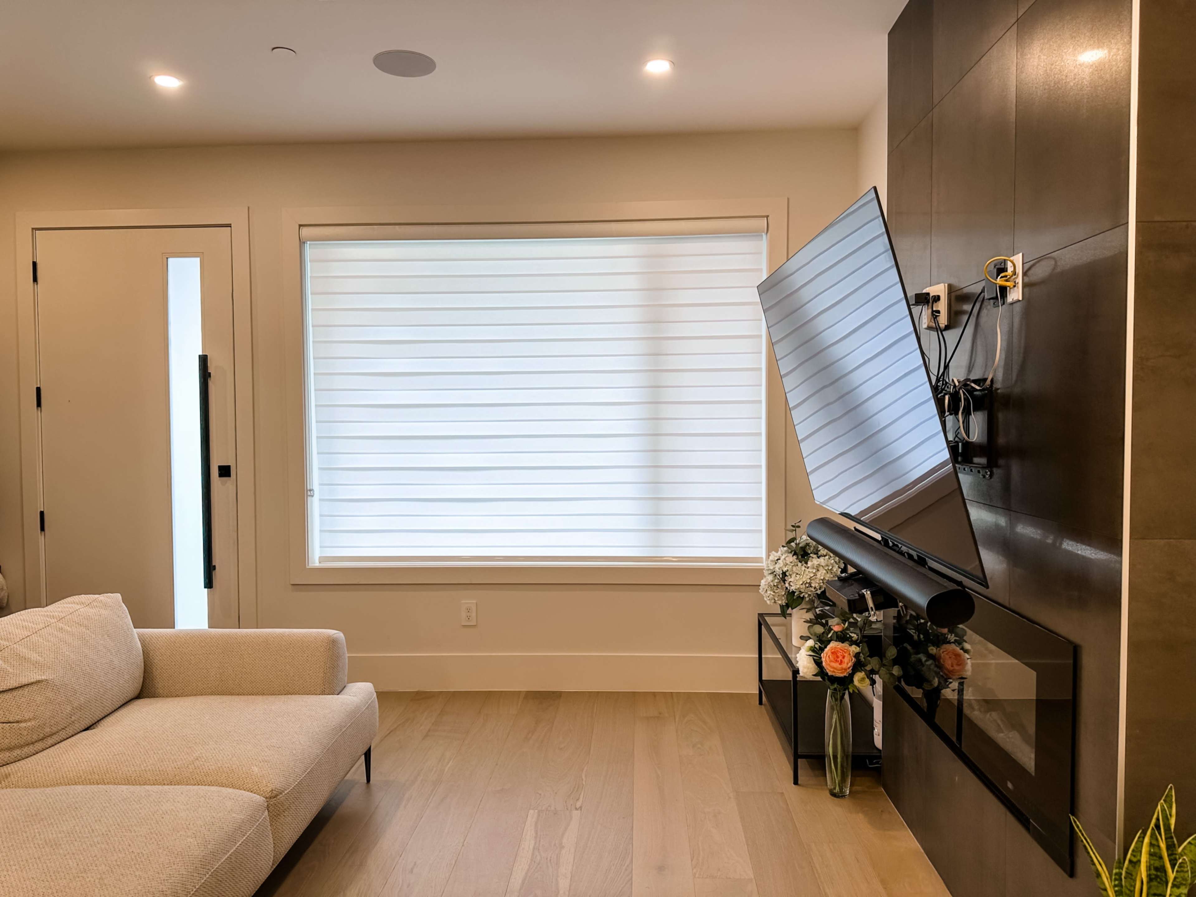 A modern living room featuring a mounted television on a tiled wall, a light-colored sofa, and a window with sheer blinds.