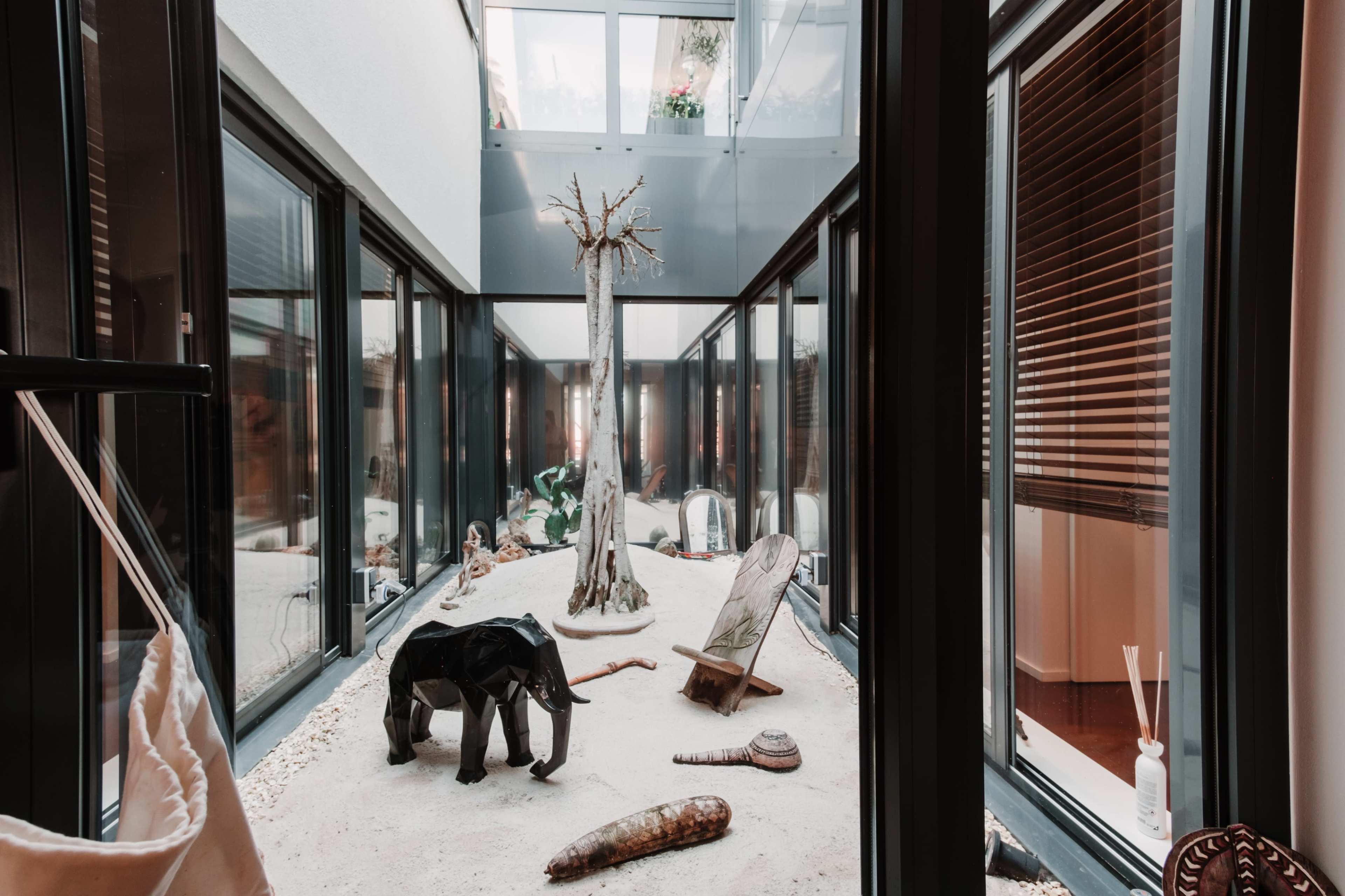 The image shows an indoor space featuring a sandy area with various decorative objects, including a black elephant sculpture and a tree, framed by glass walls.