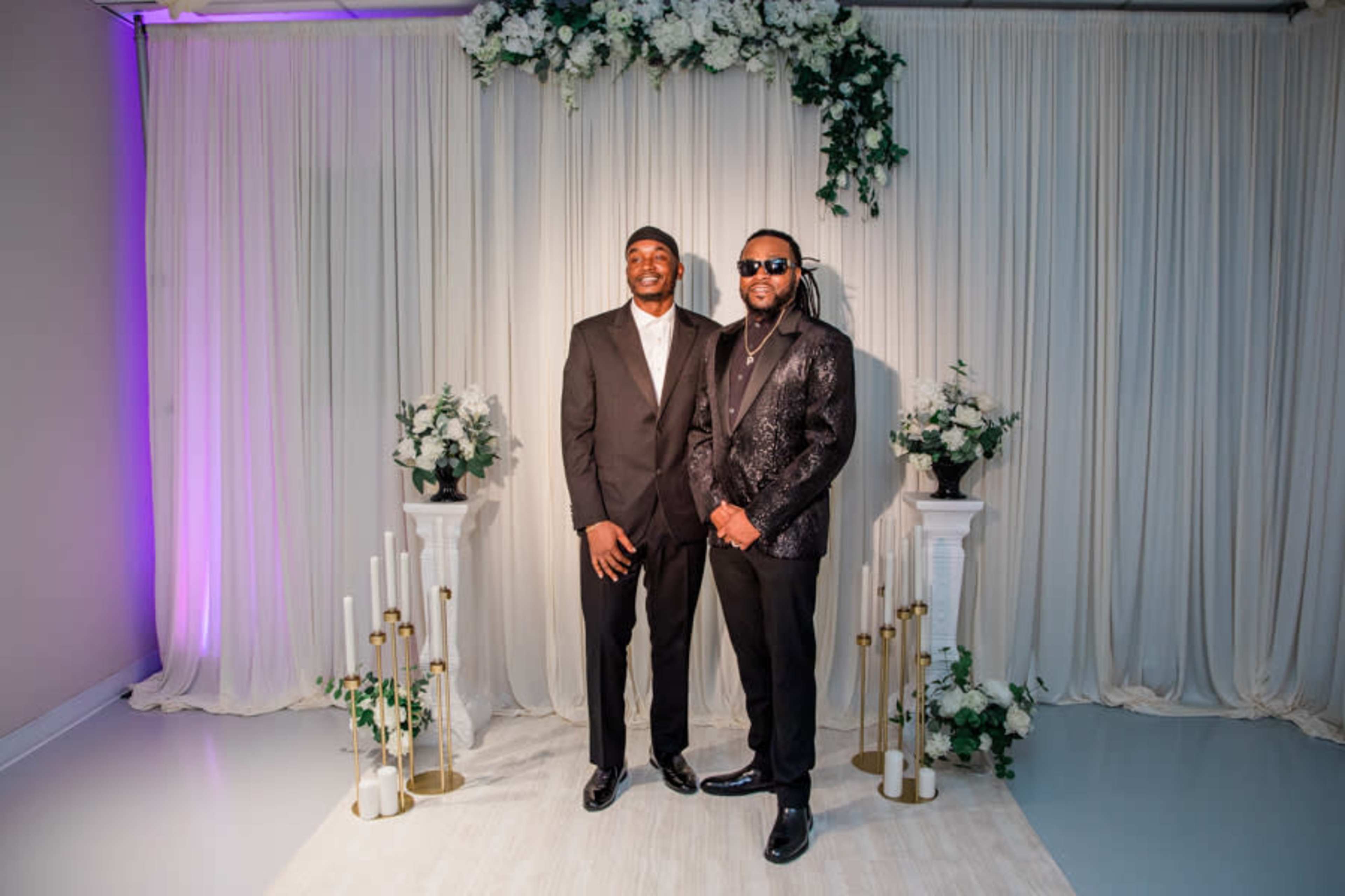 Two men stand confidently in formal attire in front of a floral-decorated backdrop with candles and greenery.
