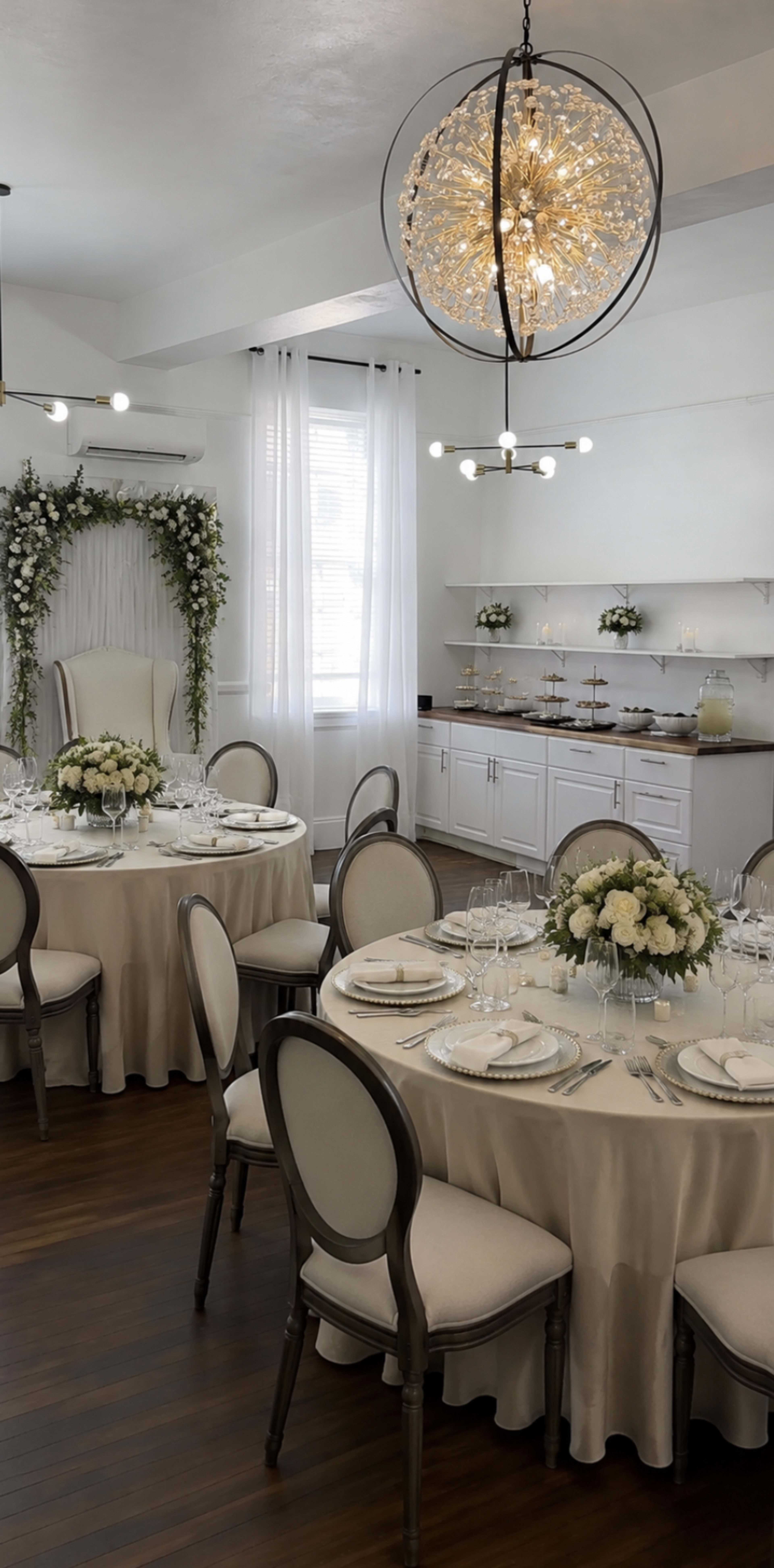 A dining room is set with elegantly arranged round tables featuring white tablecloths, fine diningware, and floral centerpieces, with decorative floral arrangements on the walls and a stylish chandelier overhead.