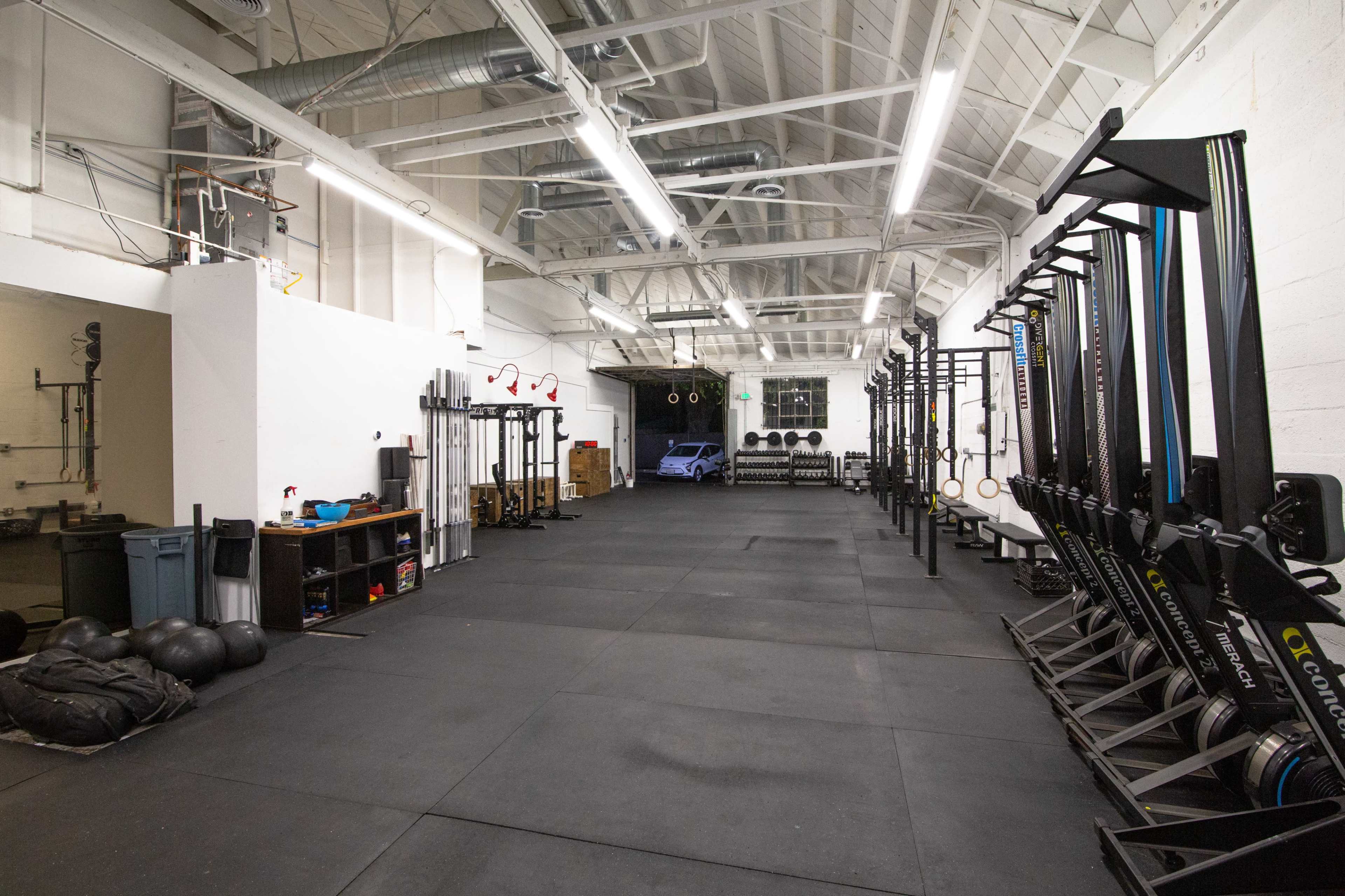 The image shows a spacious gym with various workout equipment, including rowing machines and weights, arranged along one wall, and a rubberized floor.