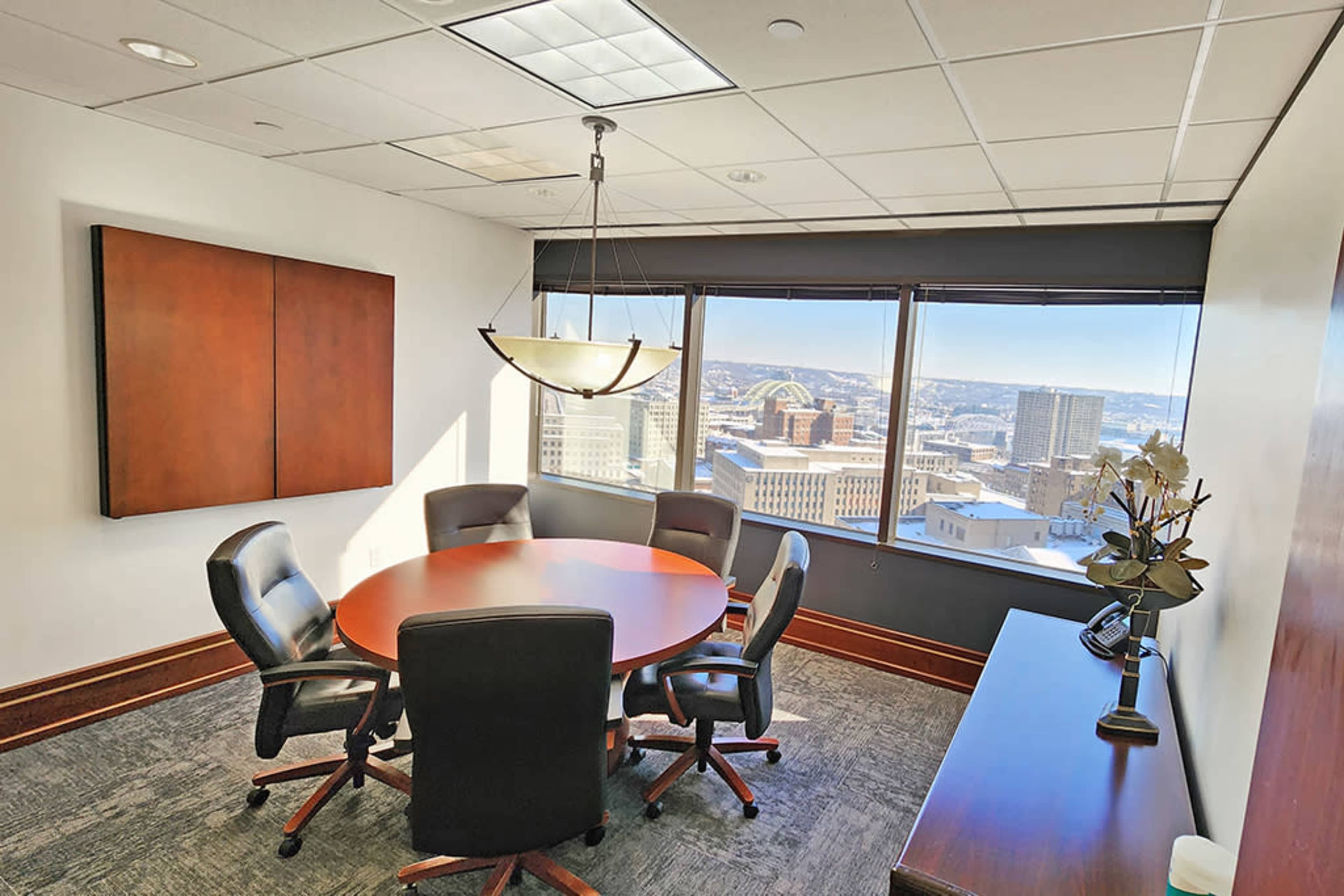 The image shows a conference room with a round table and black chairs, featuring large windows that offer a view of a city skyline.