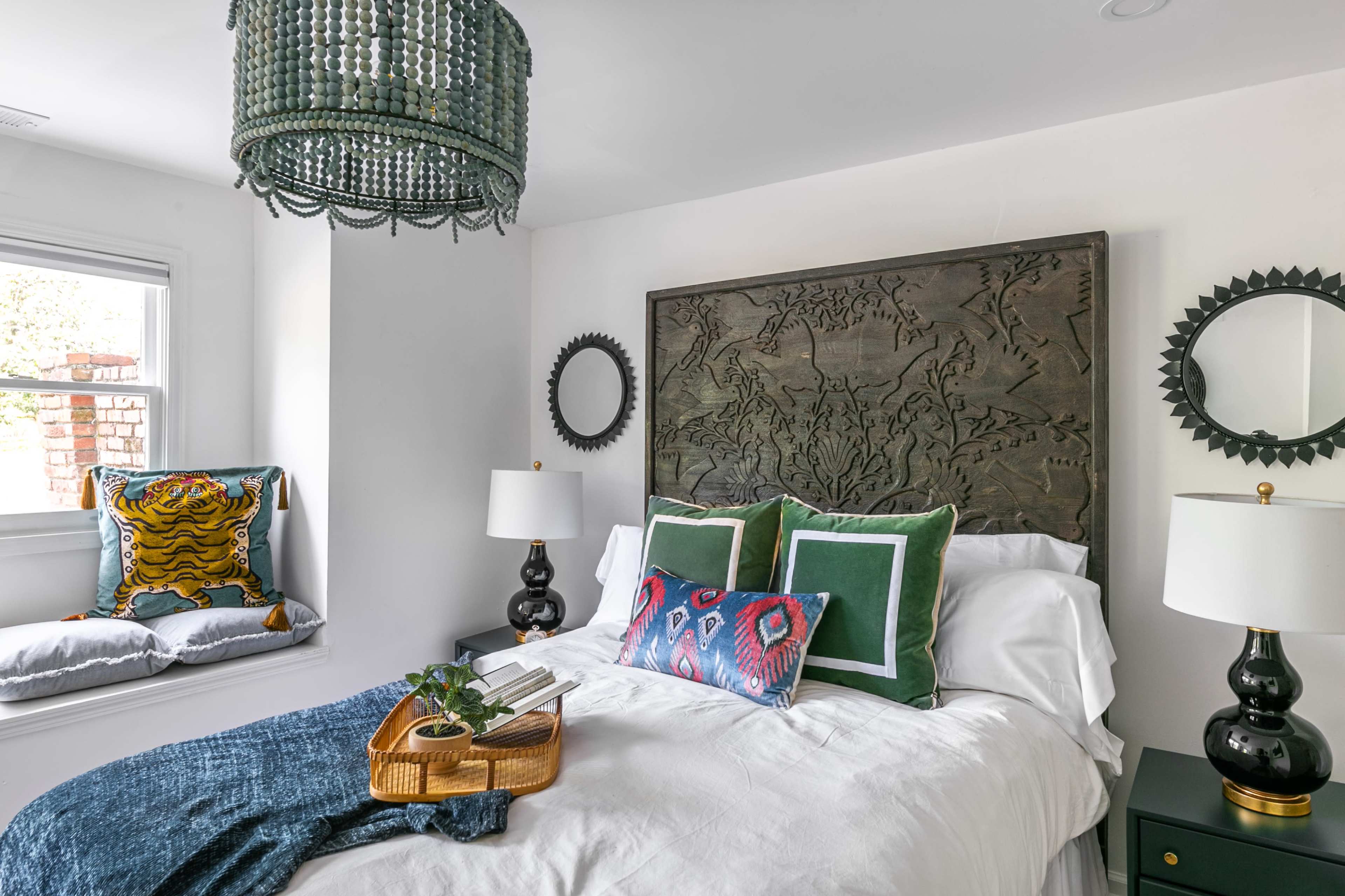 A well-decorated bedroom features a bed with decorative pillows, a textured headboard, two lamps on nightstands, and a window seat with cushions.