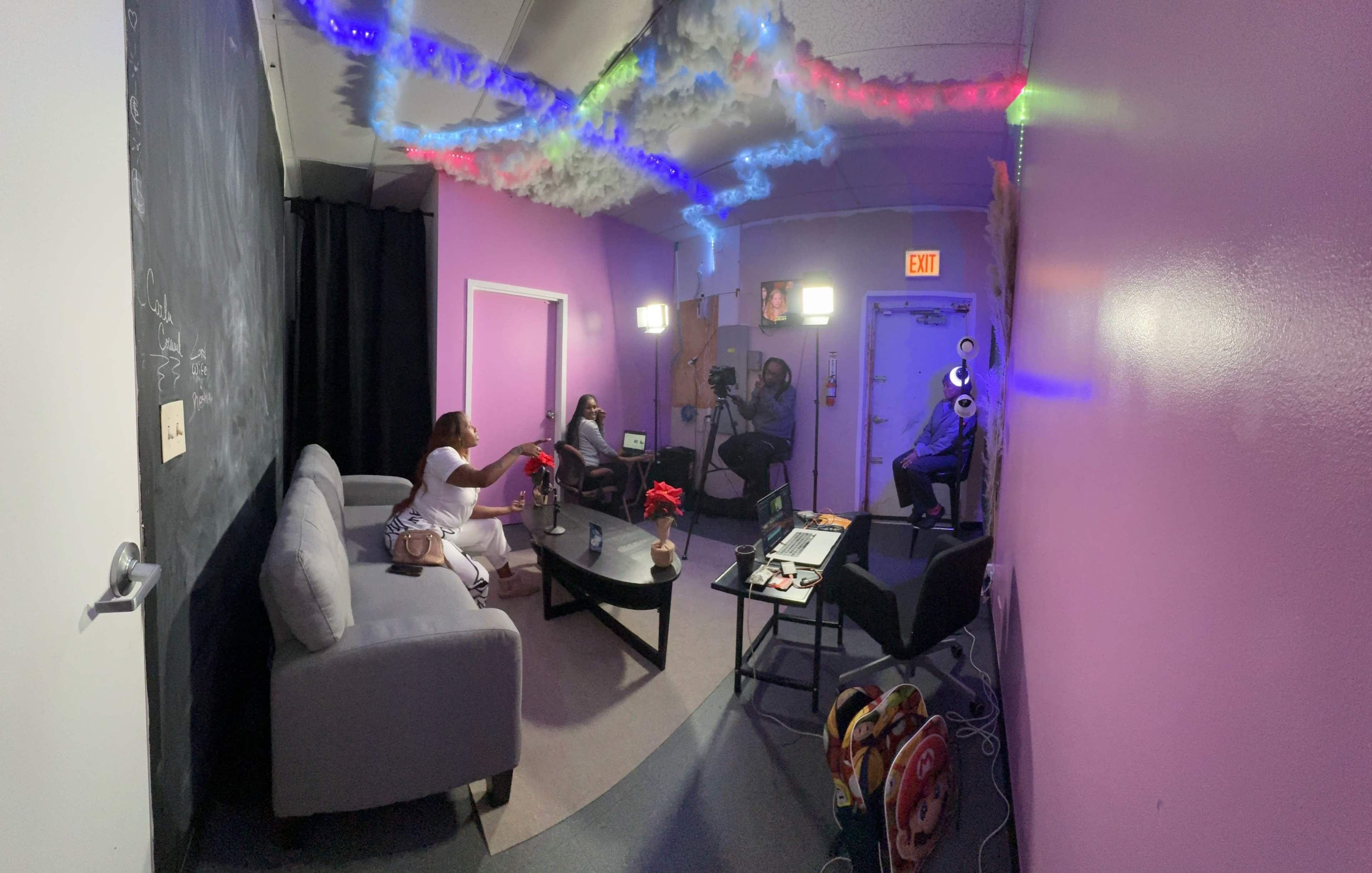 A small, colorful studio space with a pink wall, a grey couch, and lights arranged on the ceiling, where three people are engaged in a video shoot.