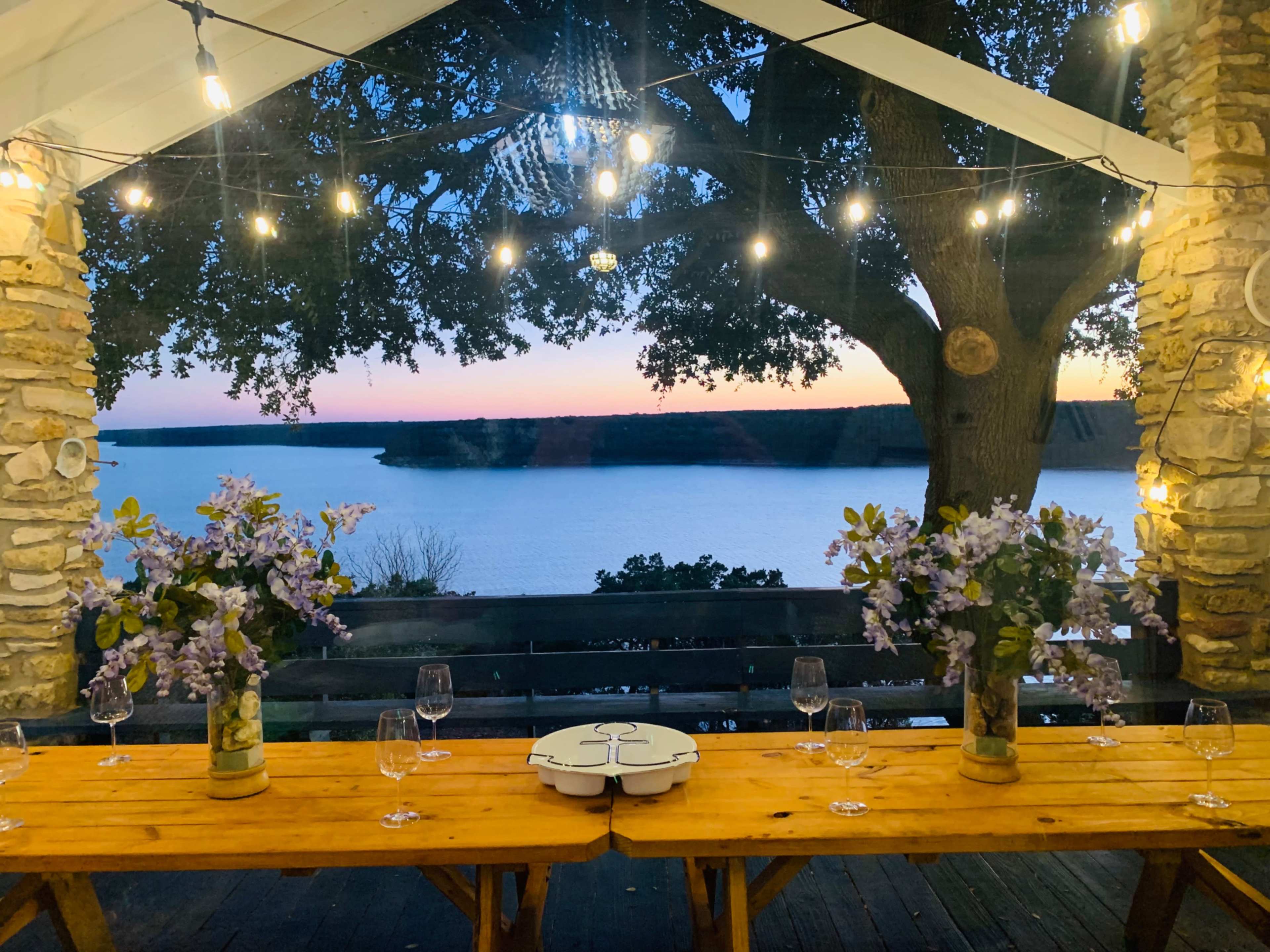 A rustic dining setup with wooden tables adorned with flower arrangements and wine glasses overlooks a calm lake at sunset, illuminated by string lights.