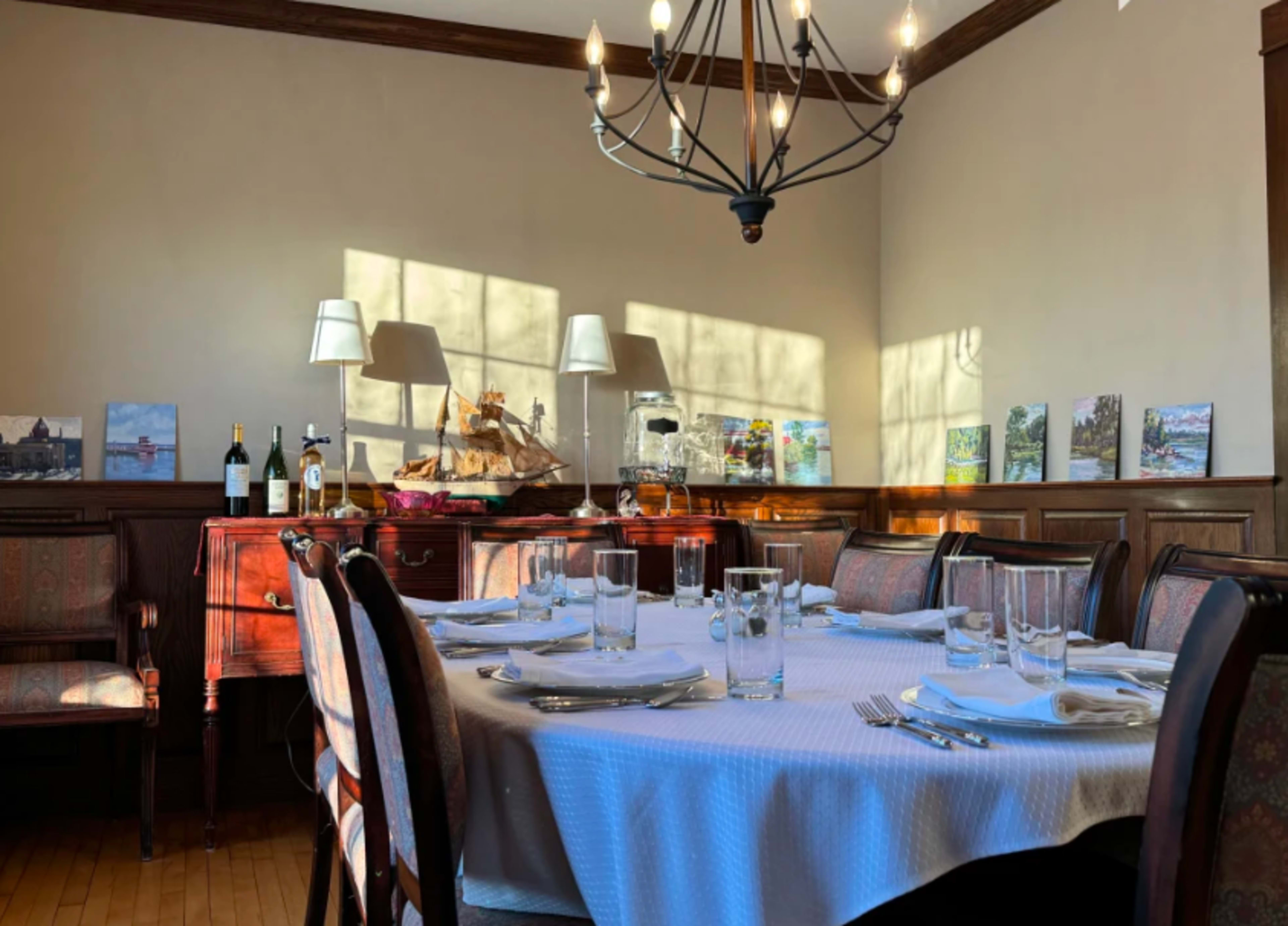 A dining room with a large round table set for a meal, surrounded by wooden chairs, and decorated with various artworks, wine bottles, and a chandelier overhead.