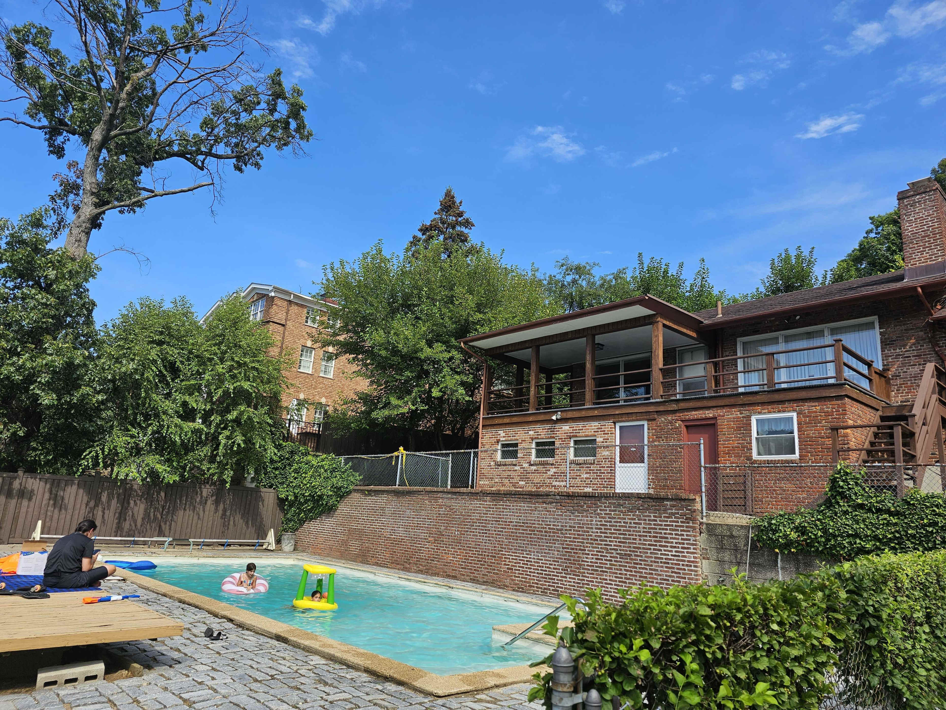 A backyard with a swimming pool surrounded by trees, a brick house with a balcony, and several people enjoying the water.
