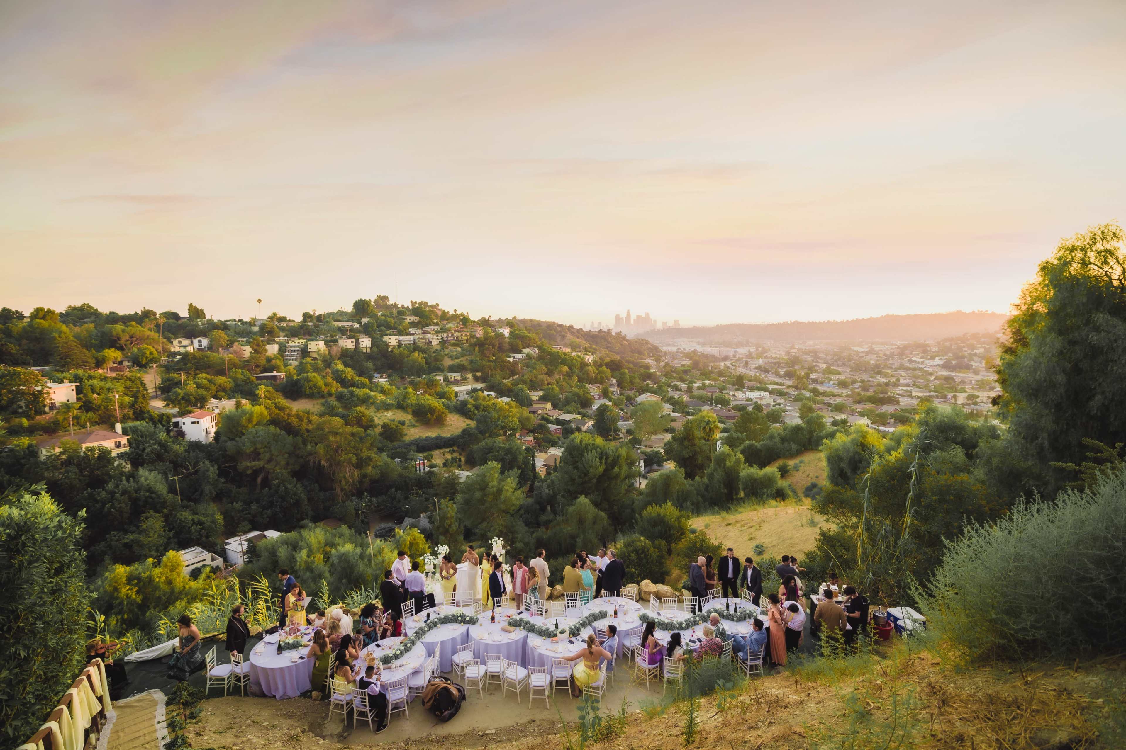 A large outdoor gathering with elegantly decorated tables is set on a hillside overlooking a city skyline at sunset.